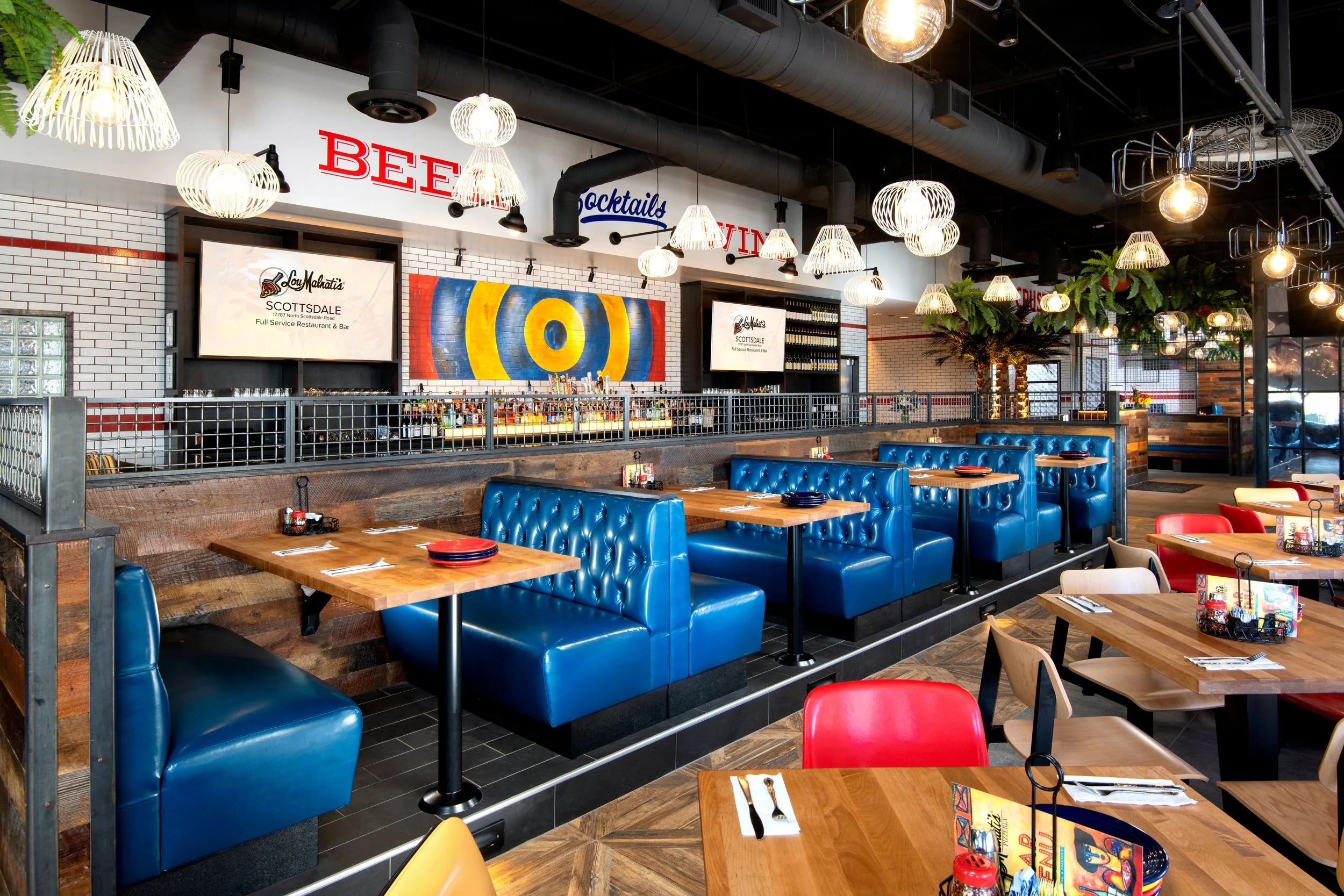 Inside a modern restaurant with blue leather booths, wooden tables, and colorful chairs. Decor includes hanging light fixtures, greenery, and wall art. The bar area has signage for beers, cocktails, and wine.