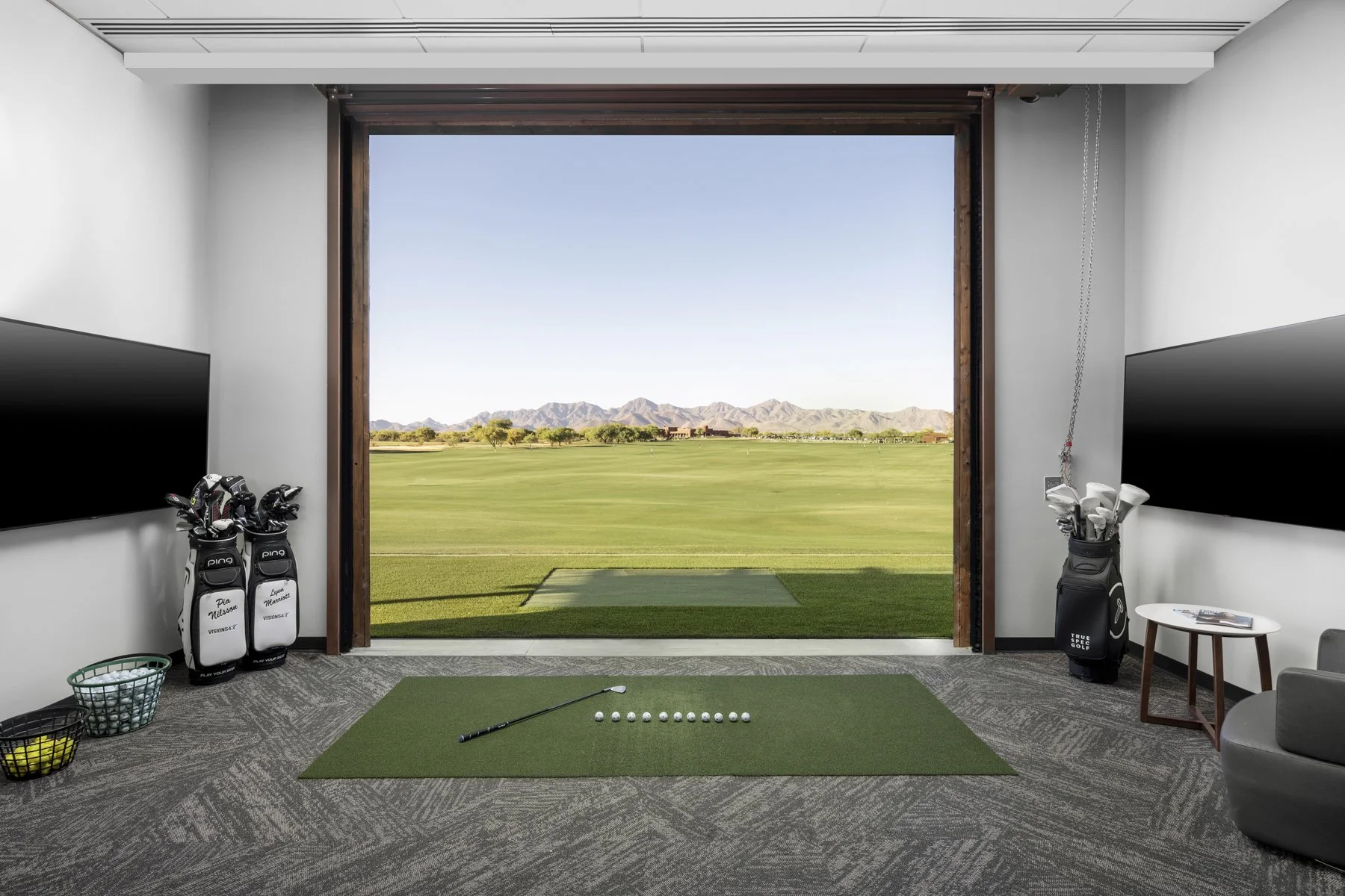 Indoor golf simulation room with a view of a golf course outside, featuring golf clubs in bags, a golf ball, and a club on a green mat.