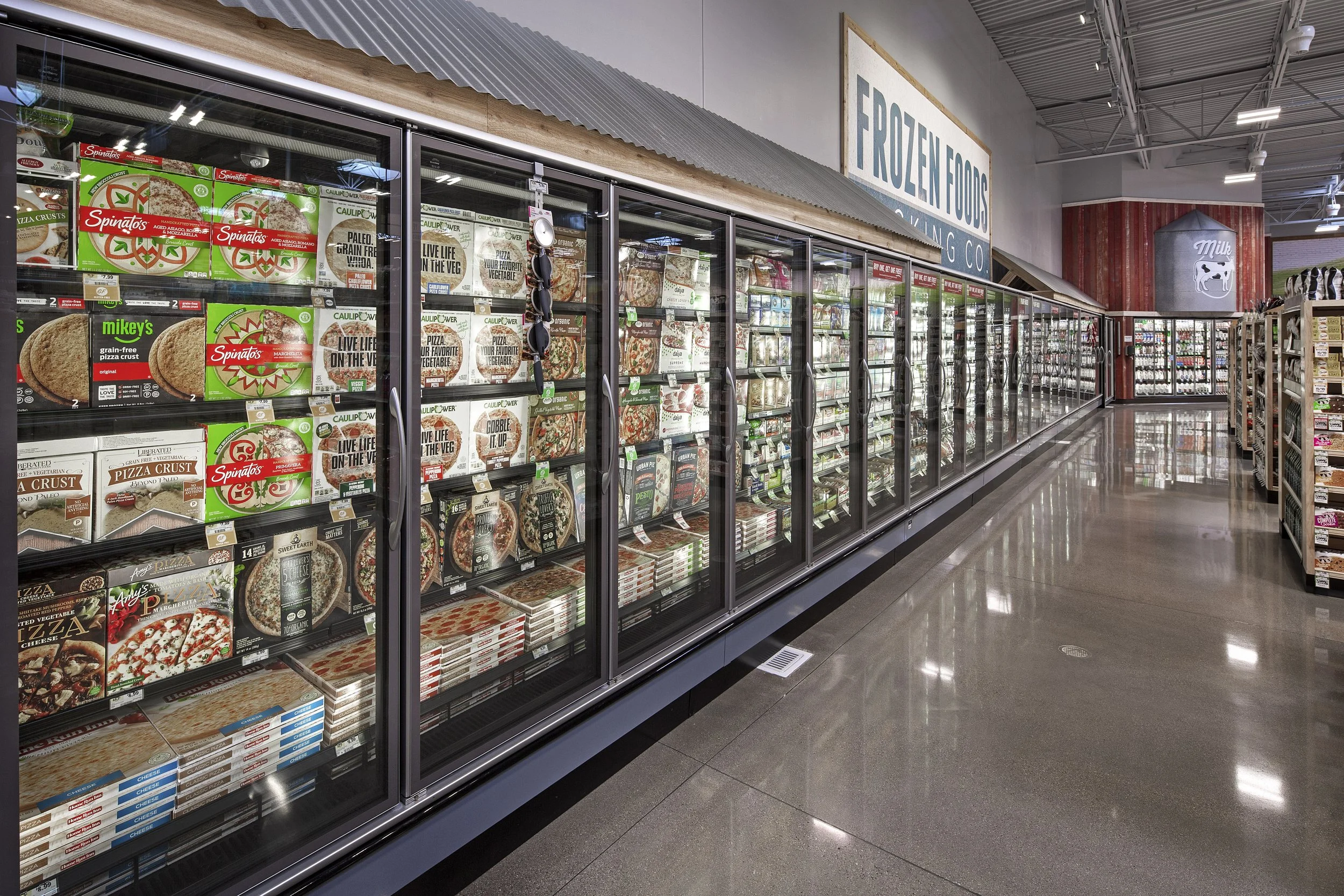 Frozen foods aisle in a grocery store with glass-door freezers filled with pizza and frozen food packages, and a large sign overhead reading 'FROZEN FOODS'.