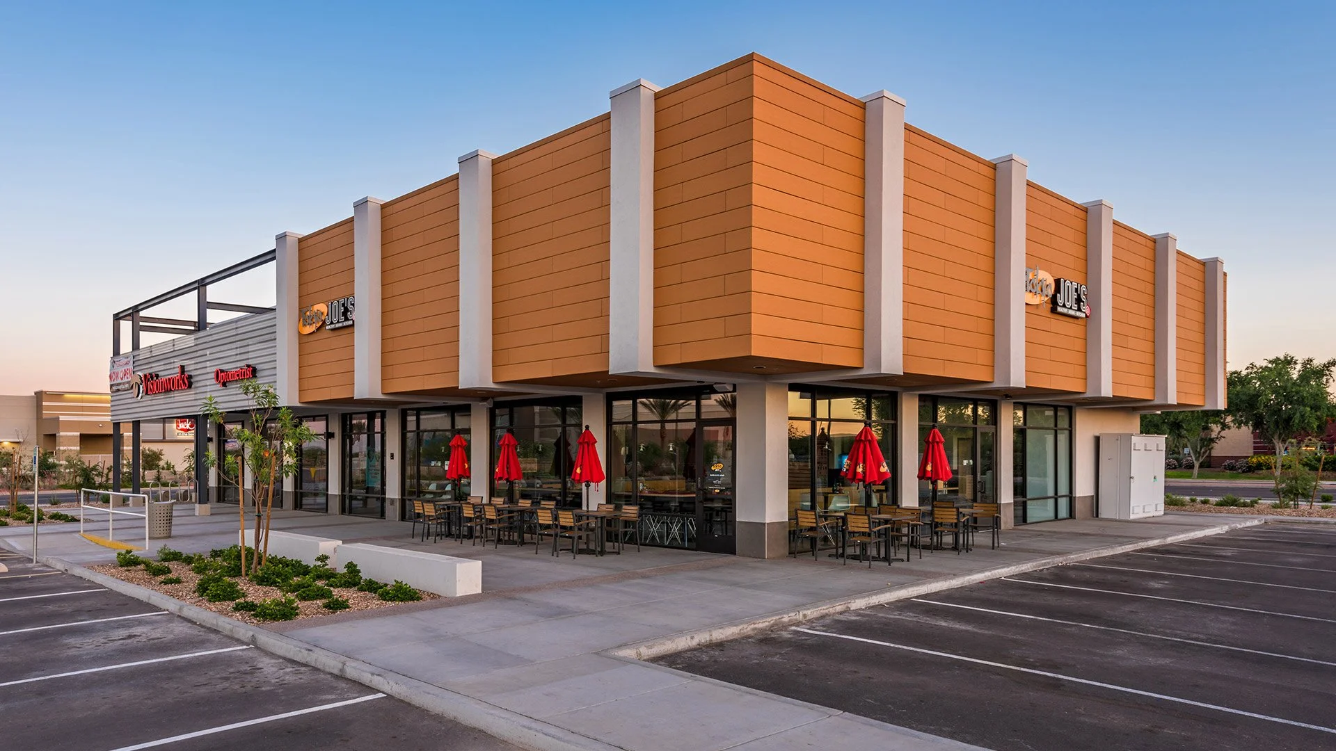 Modern commercial building with outdoor seating, red umbrellas, and empty parking lot at sunset.