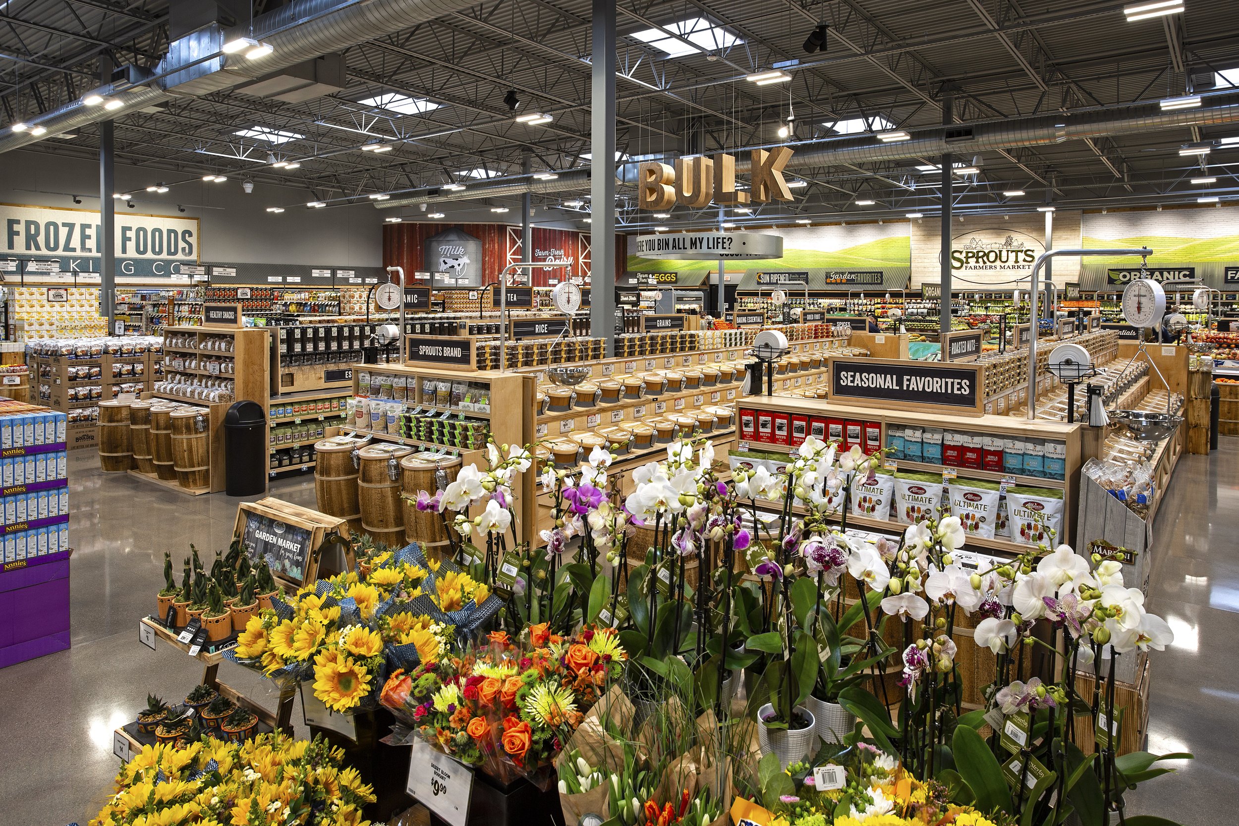 Interior of a grocery store with sections for bulk foods and seasonal favorites, shelves stocked with products, and floral displays including sunflowers and orchids.