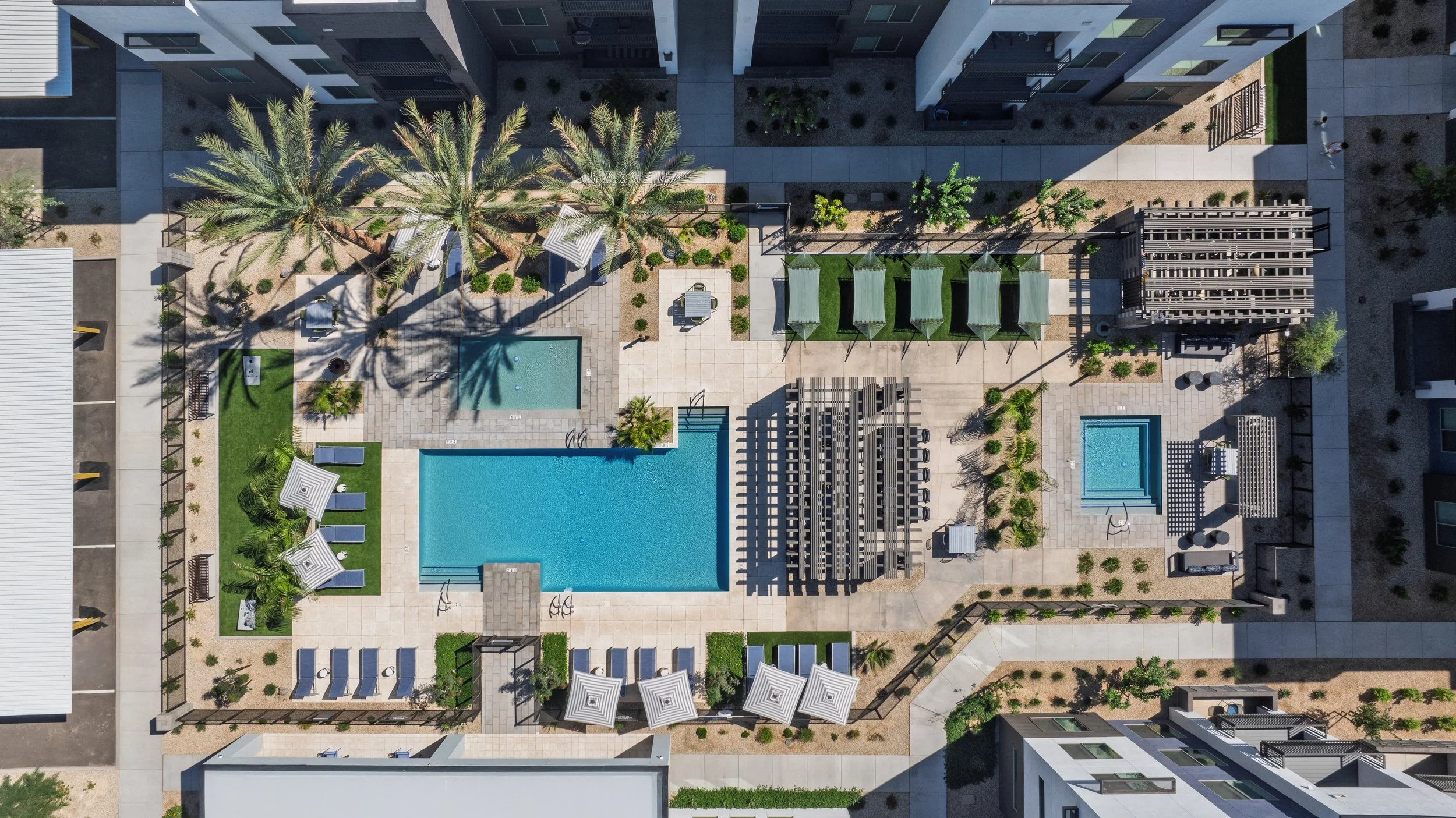 Aerial view of a modern apartment complex outdoor pool area featuring two rectangular pools, poolside lounge chairs with umbrellas, green cabanas, palm trees, a hot tub, and surrounding pathways and landscaped areas.