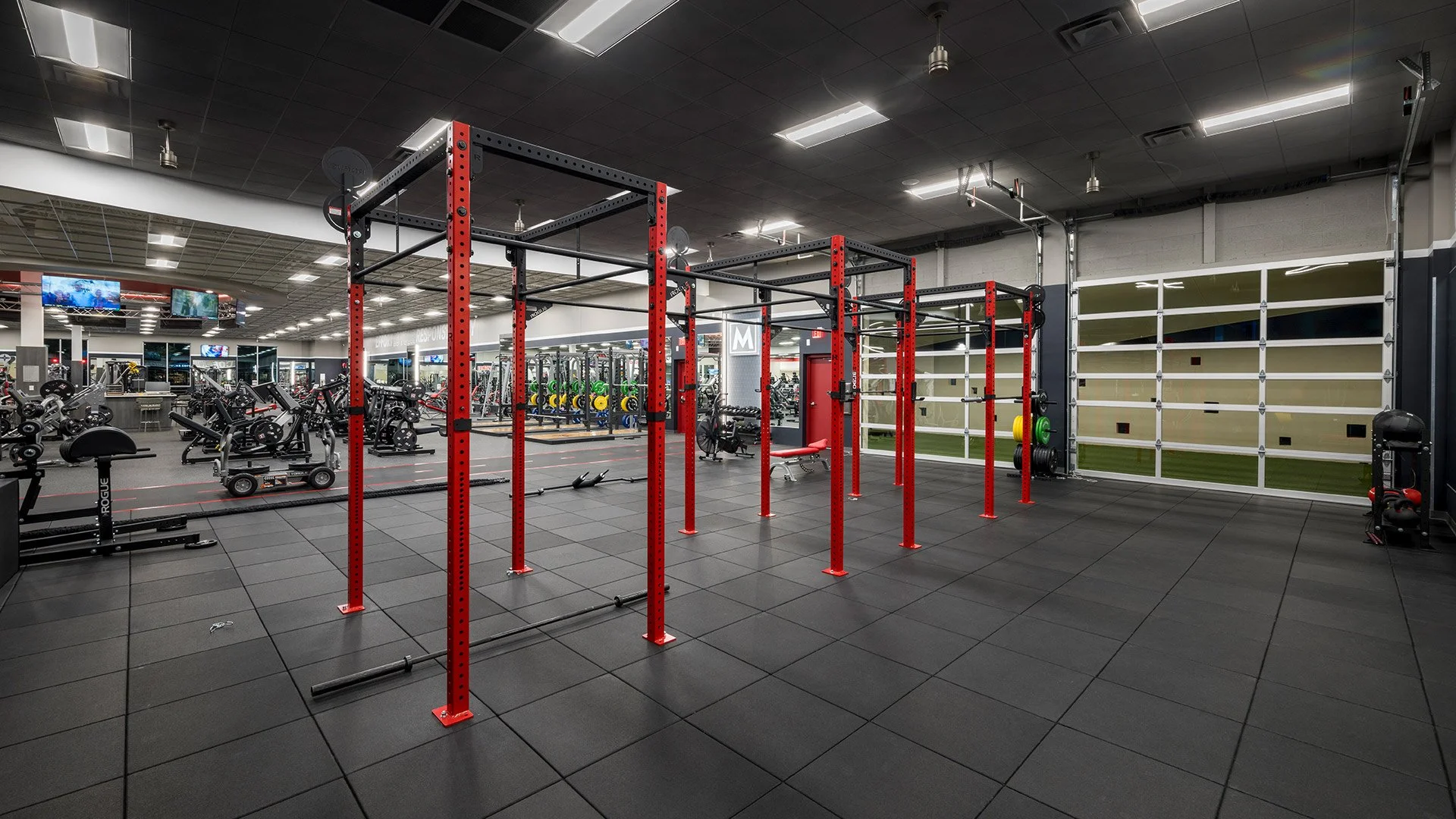 Empty gym workout area with red and black pull-up bars, black rubber flooring, and various exercise equipment in the background. The gym is well-lit with ceiling lights and has a mirrored wall on the left.