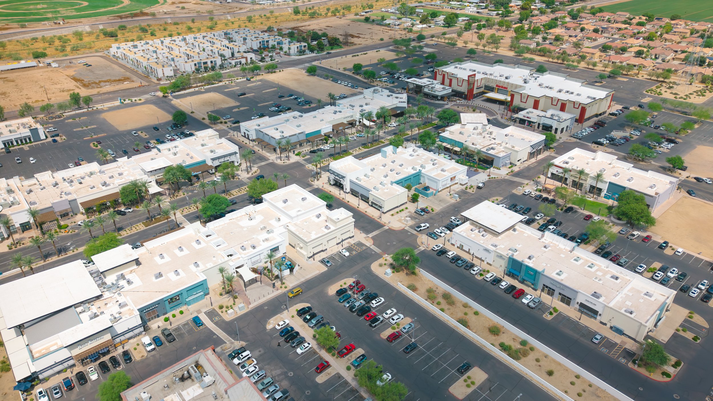 Aerial view of a commercial shopping center with retail stores, parking lots, and surrounding residential neighborhoods, including a large store with a white roof and red accents, and a nearby apartment complex.