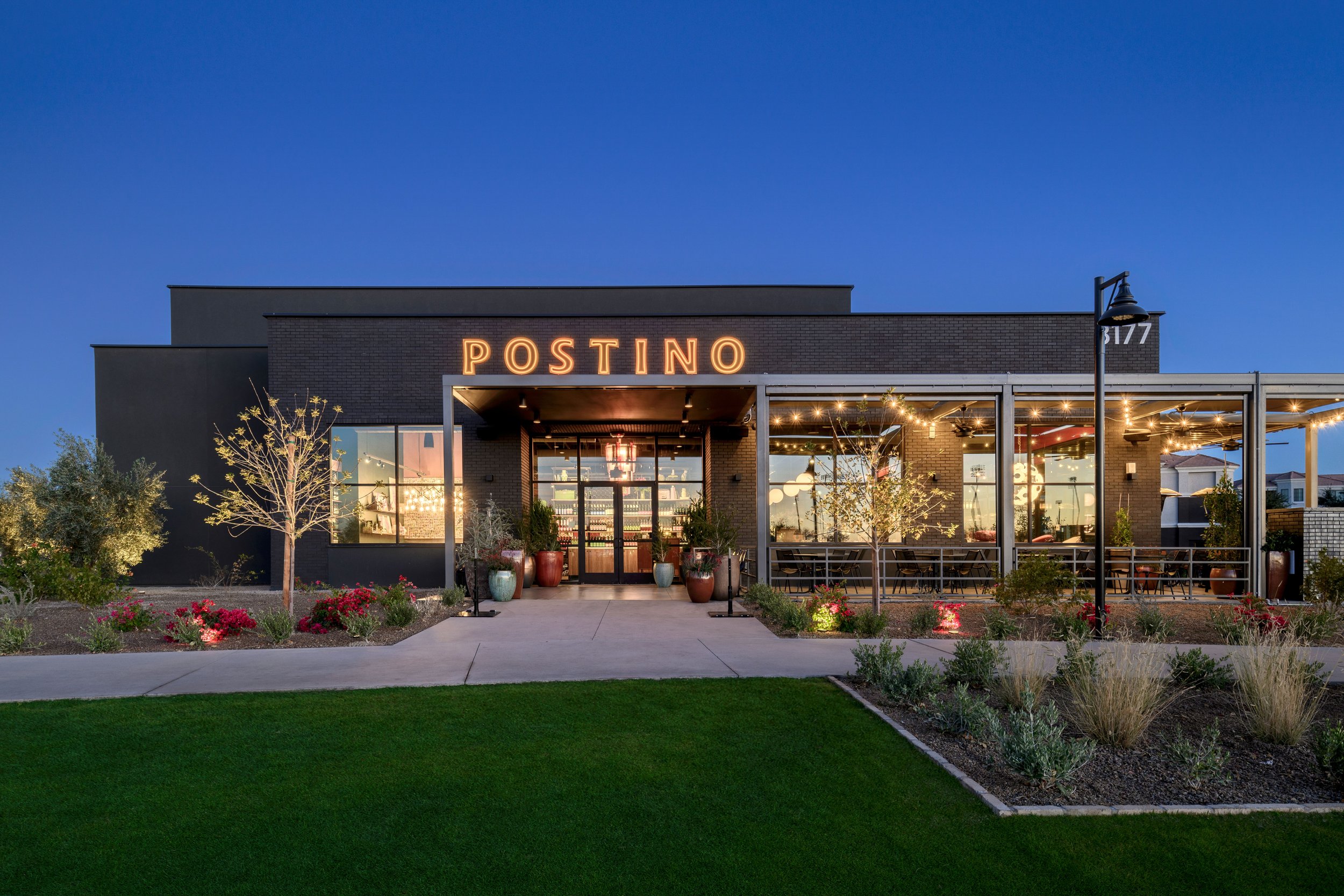 Exterior of a modern restaurant named POSTINO during evening with illuminated sign, large windows revealing the interior, landscaped garden with trees and bushes, and a sidewalk in front.