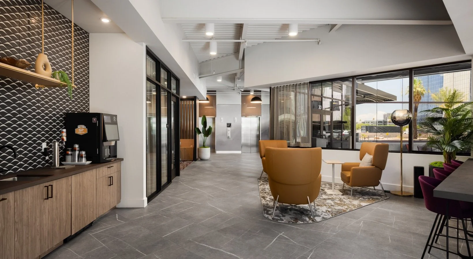 Modern hotel lobby with seating area, large windows, potted plants, elevator doors, and a coffee station with a coffee machine and cups.