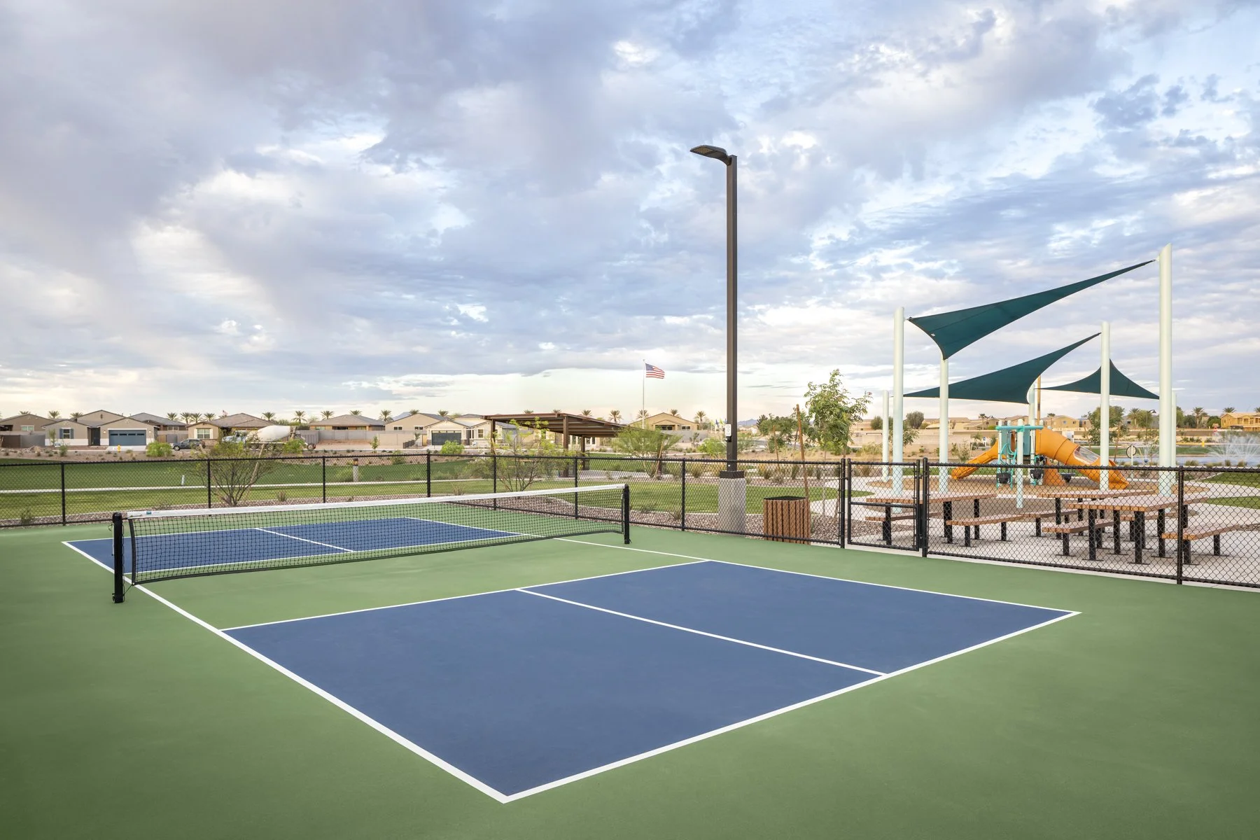 Empty outdoor tennis court with a net, surrounded by a black fence, with a playground area with shade sails and a water slide in the background, under a cloudy sky.
