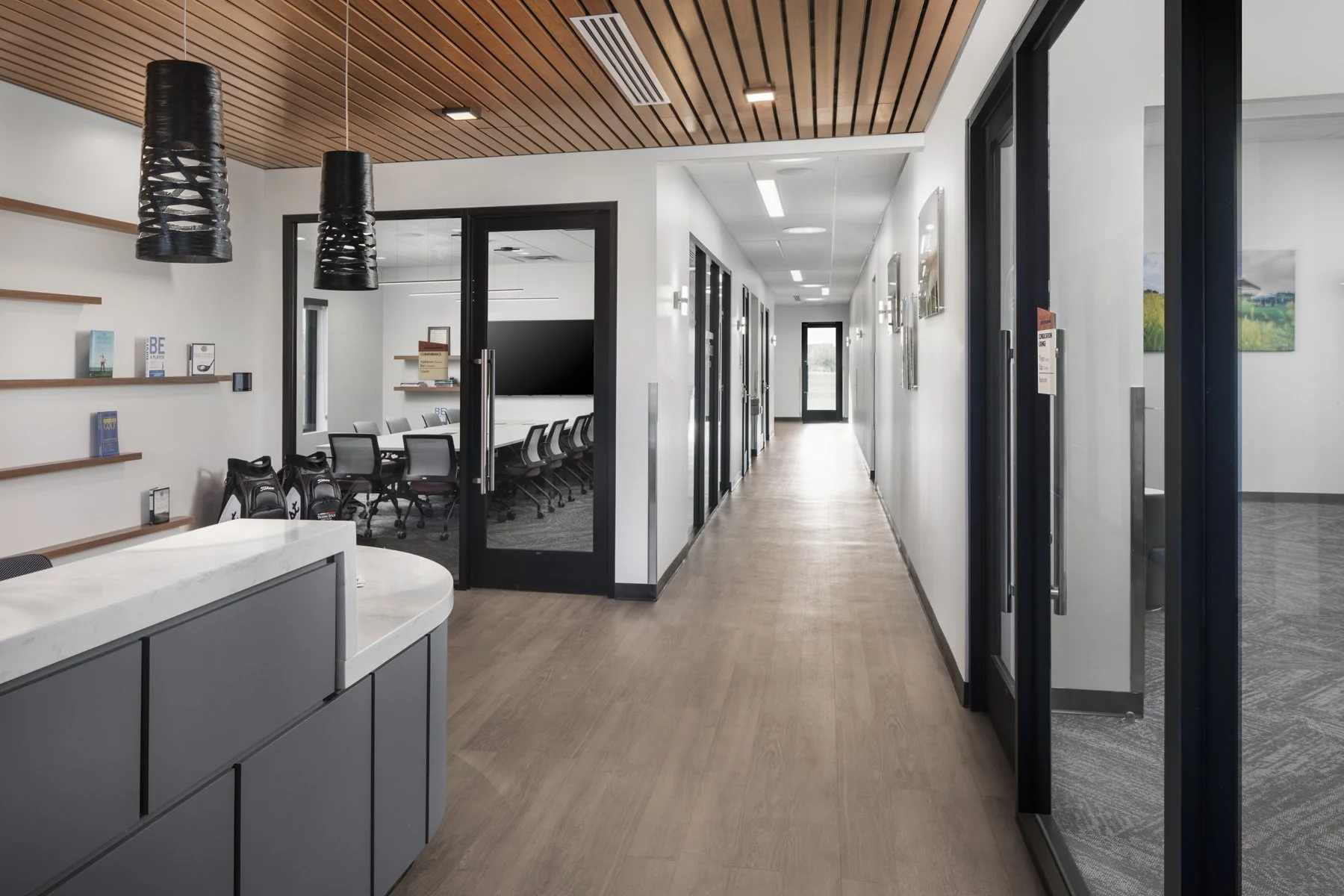 Modern office hallway with glass doors, framed pictures on white walls, wooden flooring, and meeting rooms.