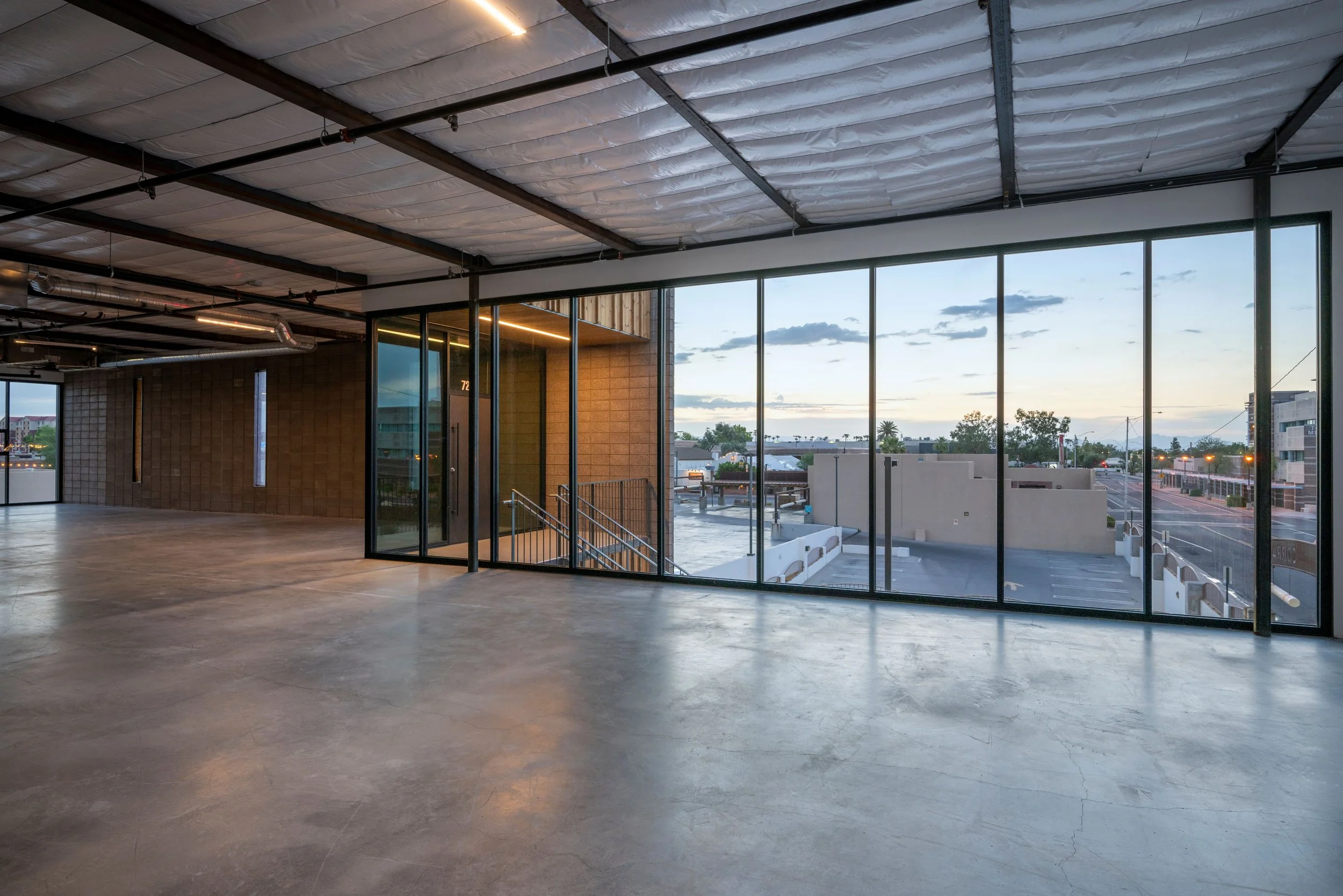 Empty interior space with concrete floor, large glass windows showing an outside view of parking lot and sky, exposed ceiling with insulation and ductwork, and an entrance with stairs outside.
