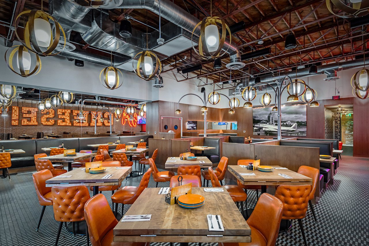 Interior of a modern restaurant with wooden tables set with plates, flatware, and napkins. Orange tufted chairs surround the tables. Decorative hanging lights and exposed ductwork are visible on the ceiling. A large illuminated sign spelling 'CRUST' 