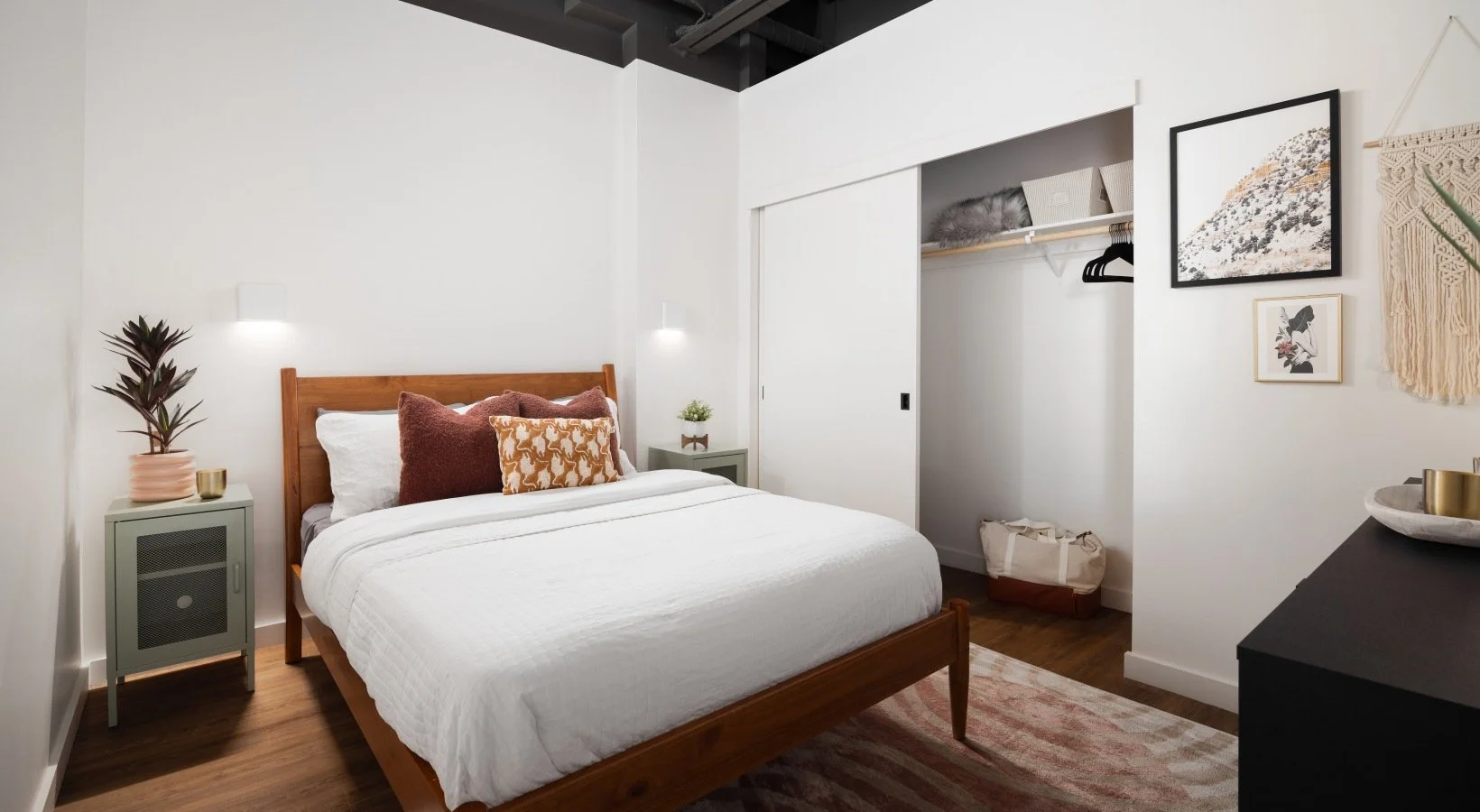A modern bedroom with a wooden bed frame, white bedding, and decorative pillows. There are small nightstands on each side with plants and decor. A closet with sliding doors is open, showing hanging clothes and storage boxes. Artwork and a wall hangin