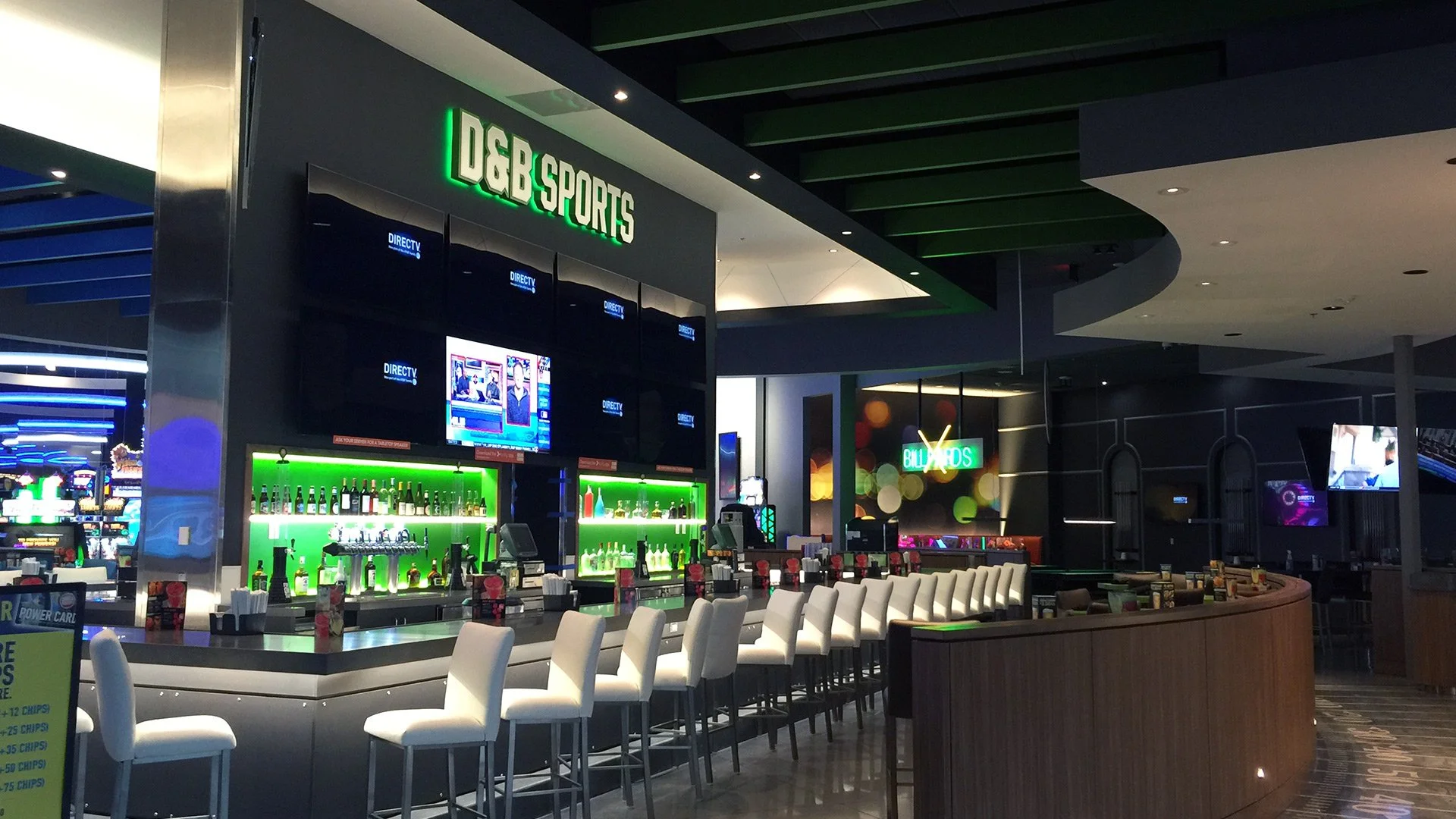 Interior of a sports bar named DGB Sports with a bar counter and row of white chairs, multiple TV screens displaying TV programs, and a smiling bartender behind the bar. Bright neon signs and modern decor enhance the ambiance.