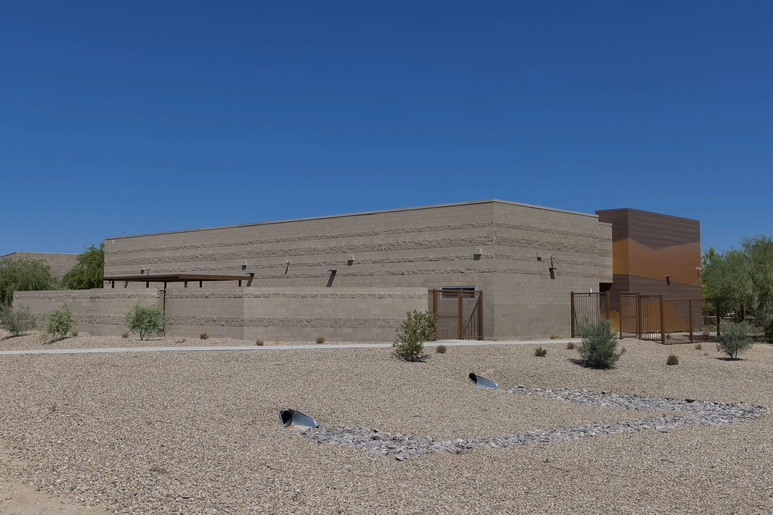 A modern building with beige and brown exterior walls, small trees and shrubs in the gravel yard, and a clear blue sky.