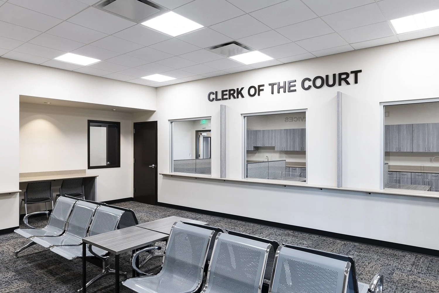 Interior of a courthouse waiting area with metal chairs, a small table, black door, and a glass windowed clerk of the court office.