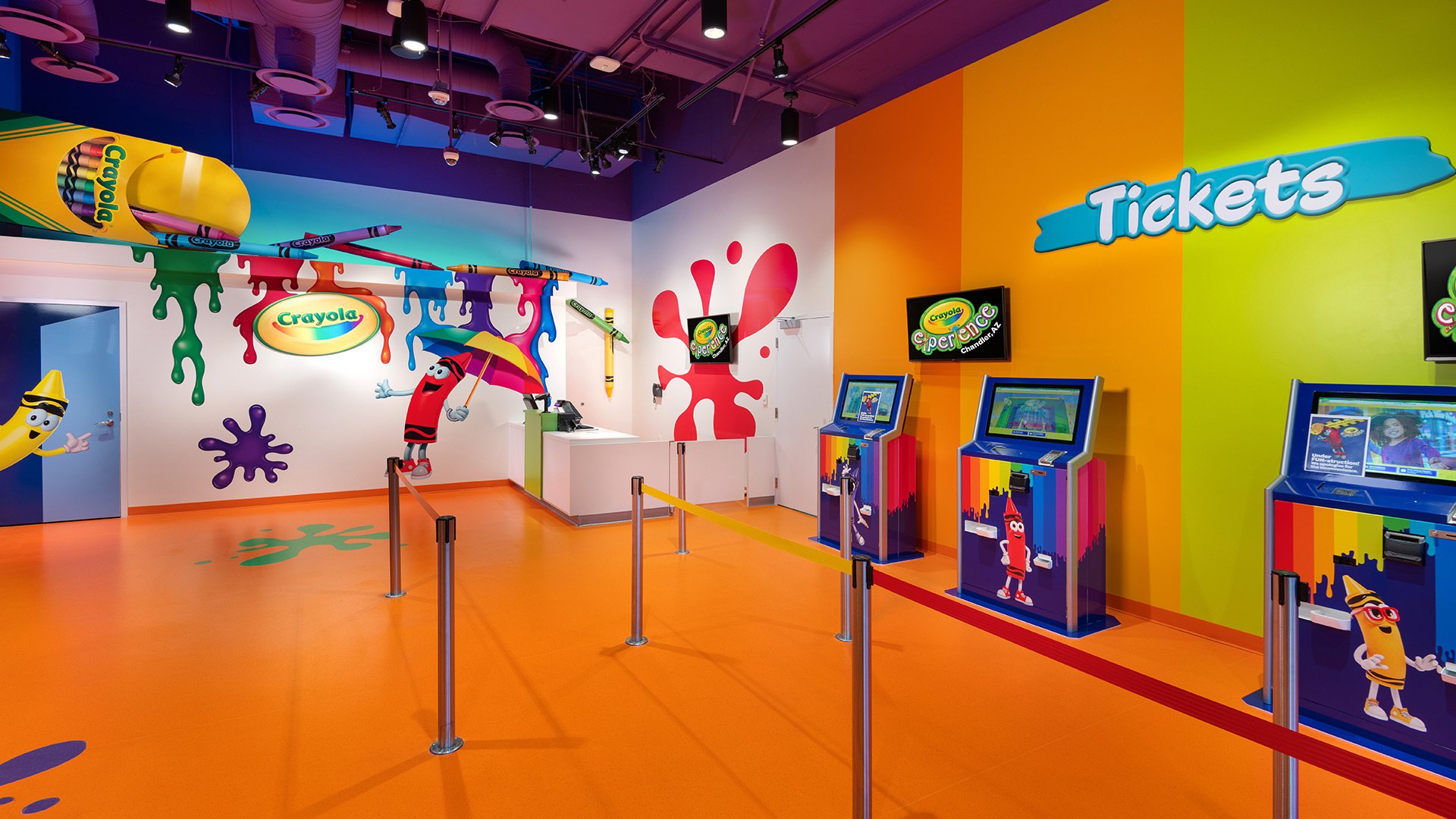Colorful indoor children’s arcade area with Crayola theme, featuring rainbow-colored walls, floor, and arcade machines, with cartoon Crayola characters, and signs reading "Tickets" and "Crayola Experience."