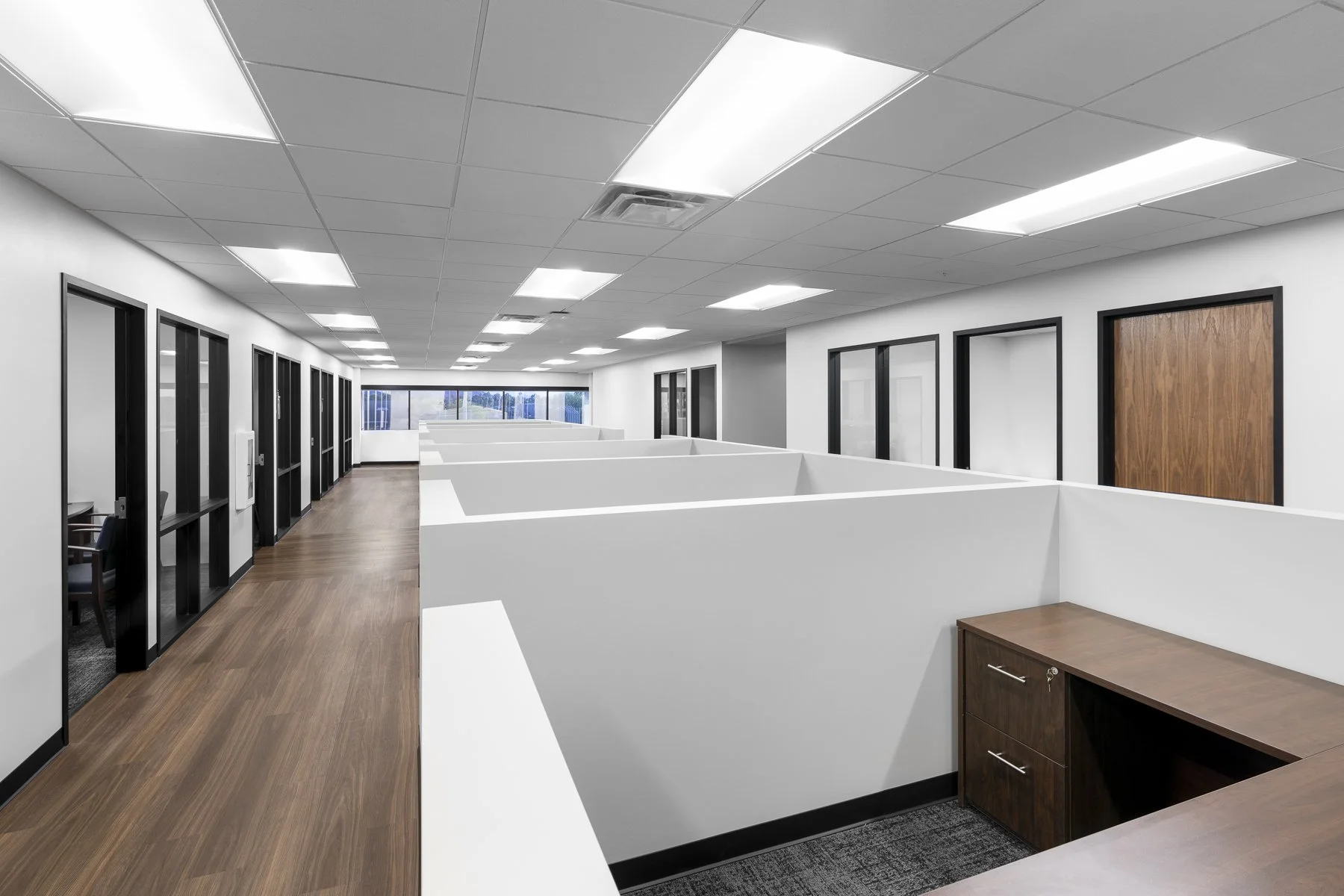 Empty office cubicles with white partitions, ceiling lights, and wooden flooring, with glass office doors on one side and a large window at the end.