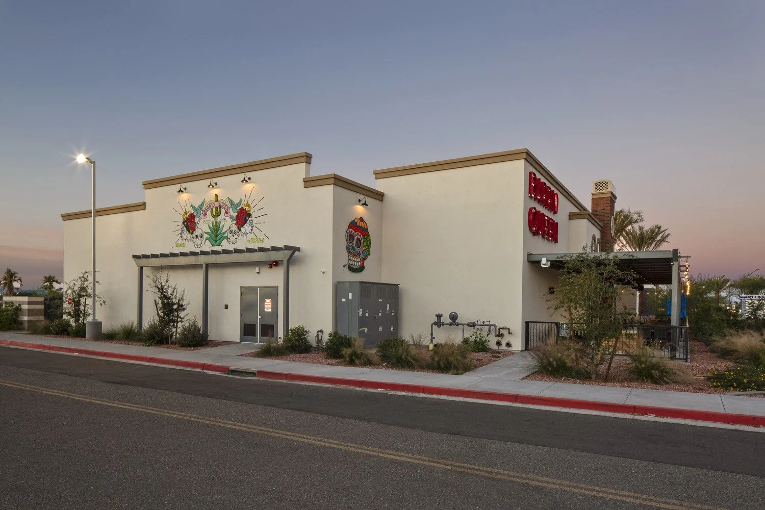 A modern building with colorful Día de los Muertos artwork on the exterior wall, including a sugar skull and decorative designs, during sunset in an urban area with desert landscaping.