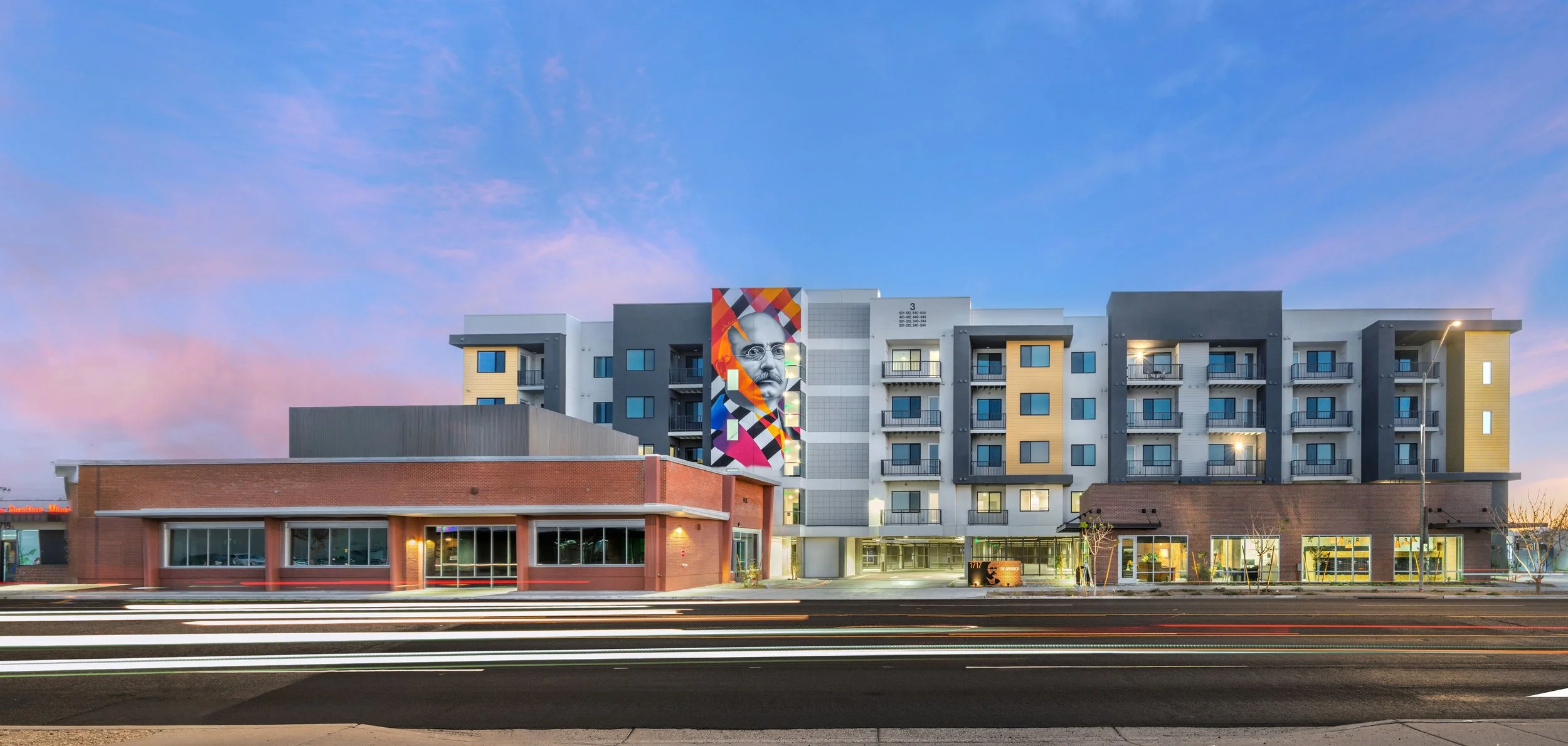 A modern multi-story apartment building with a colorful mural of a man's face on the side, taken during dusk with a pink and blue sky in the background.
