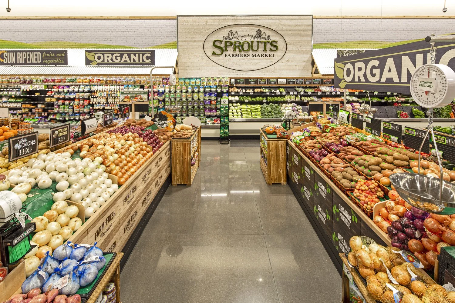 Interior of a grocery store with produce sections featuring onions, sweet potatoes, and other vegetables, with signs indicating organic and sun-ripened fruits and vegetables.