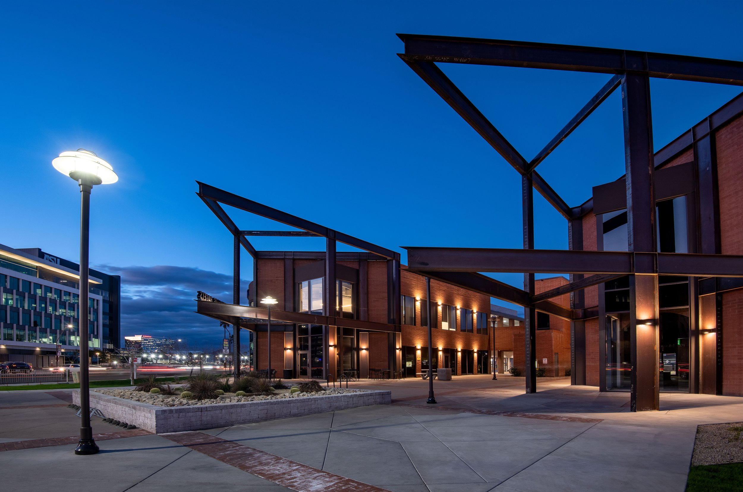 Modern building with brick walls, large windows, and metal awnings at dusk, illuminated by outdoor lights and a streetlamp, with a landscaped area and a cityscape in the background.