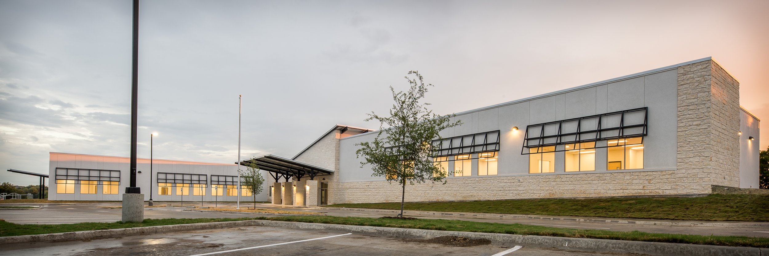 Modern commercial building with large windows, surrounded by parking lot and trees at dusk.