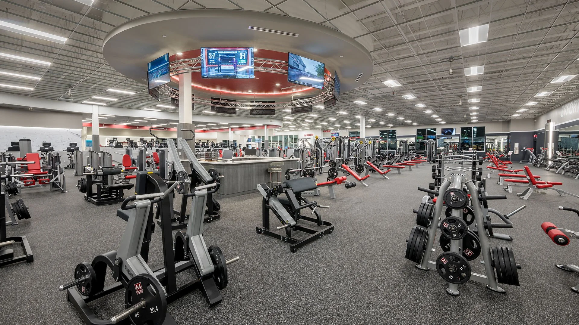 Inside view of a modern gym with various exercise equipment, cardio machines, weightlifting stations, large screens hanging from the ceiling, and bright lighting.