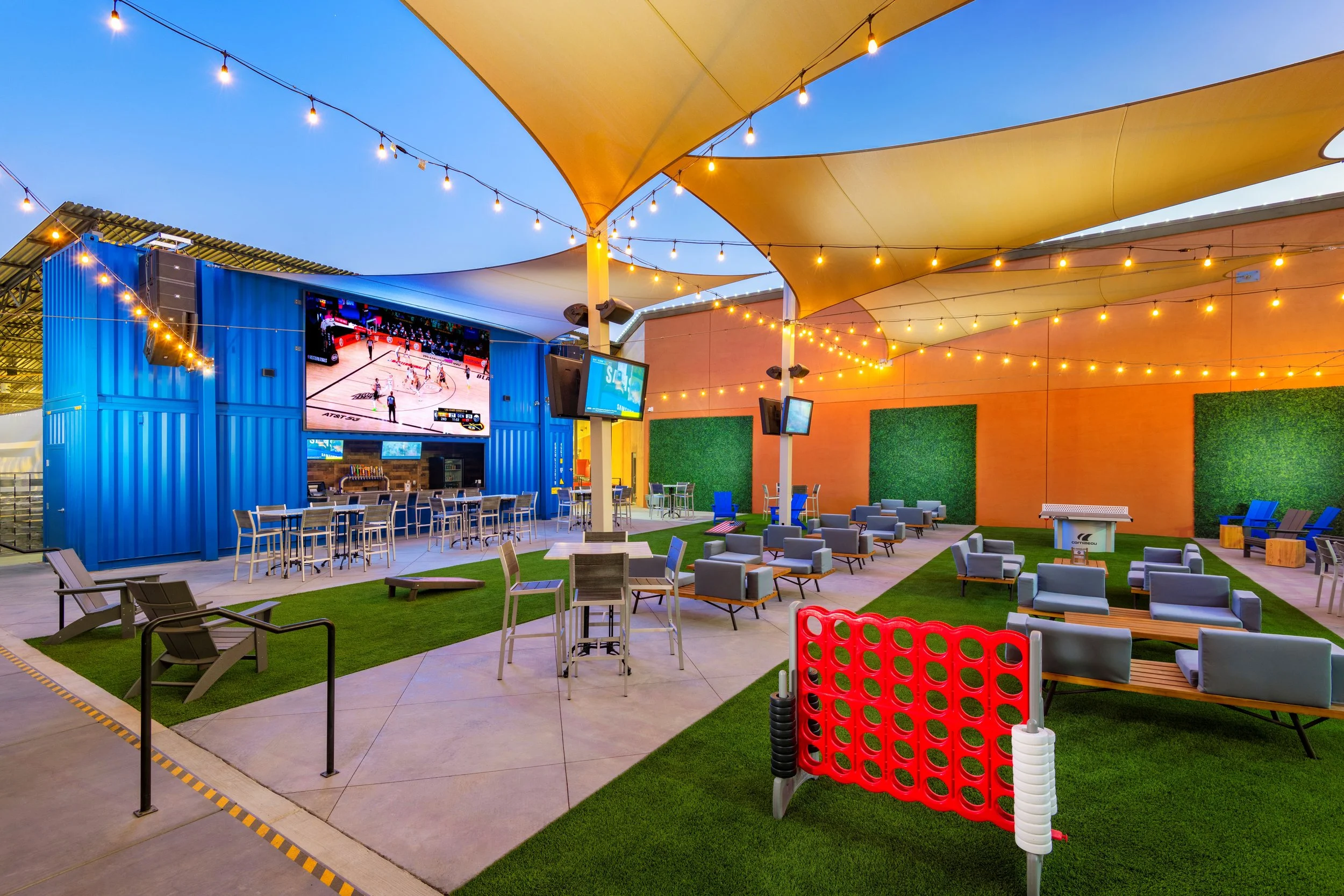 Outdoor lounge area with string lights, multiple TVs, and a large screen showing a basketball game, with blue shipping container as a bar and green decorative walls.