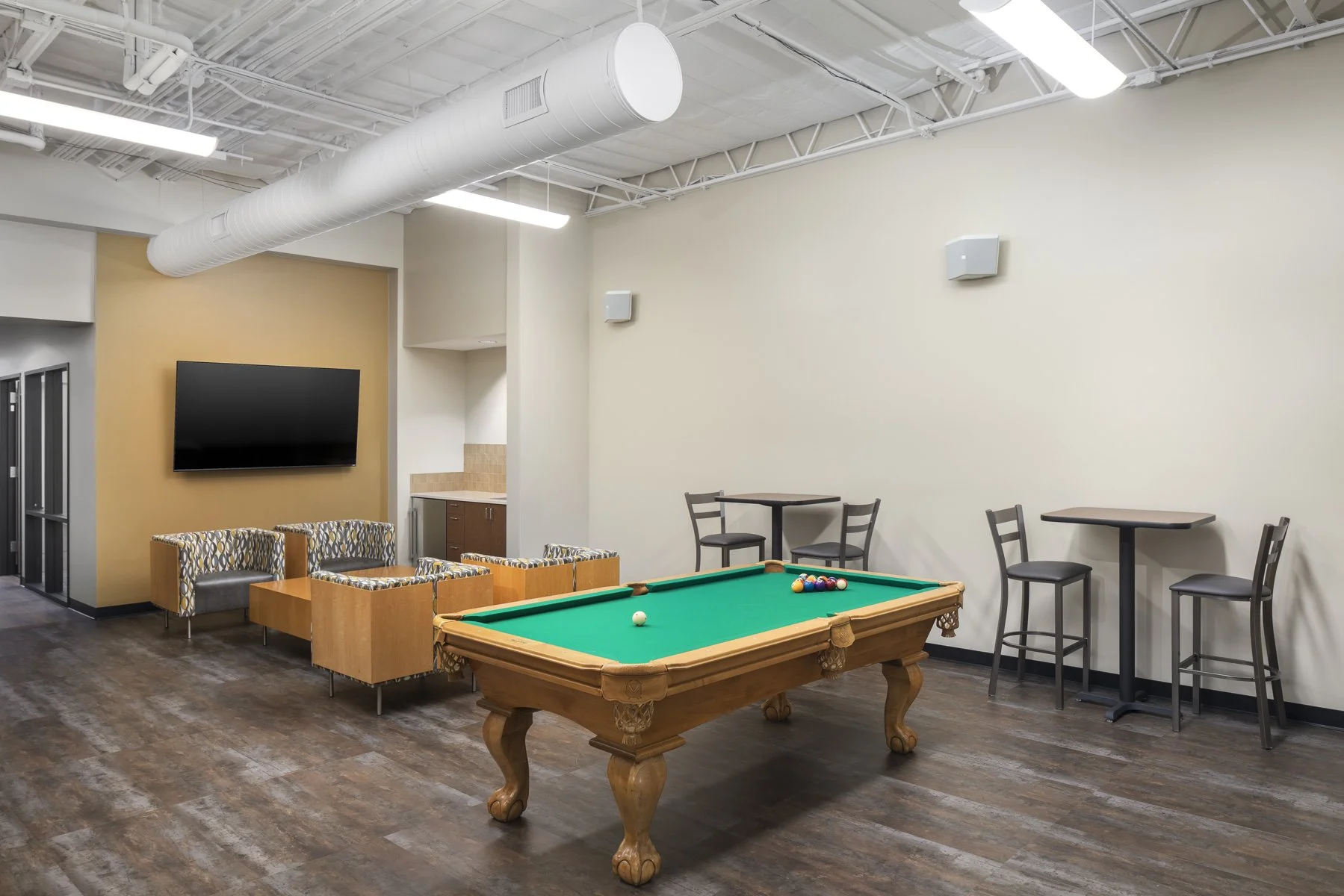 An indoor recreation room with a pool table, chairs, small tables, a television, and a small kitchenette in the background.