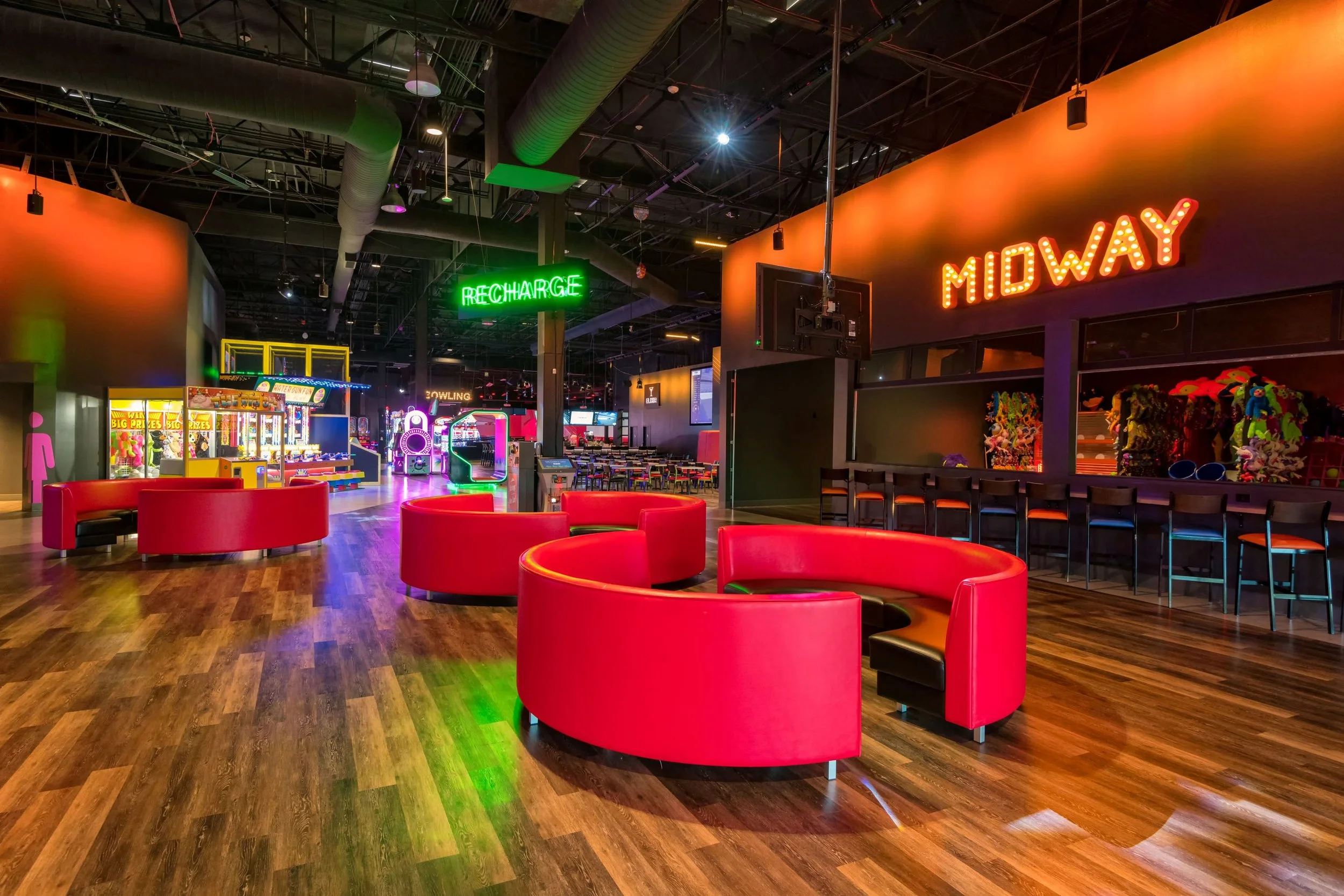 An indoor arcade with red booths, arcade game machines, and neon signs, including 'RECHARGE' in green and 'MIDWAY' in orange, with wooden flooring and a dark ceiling.