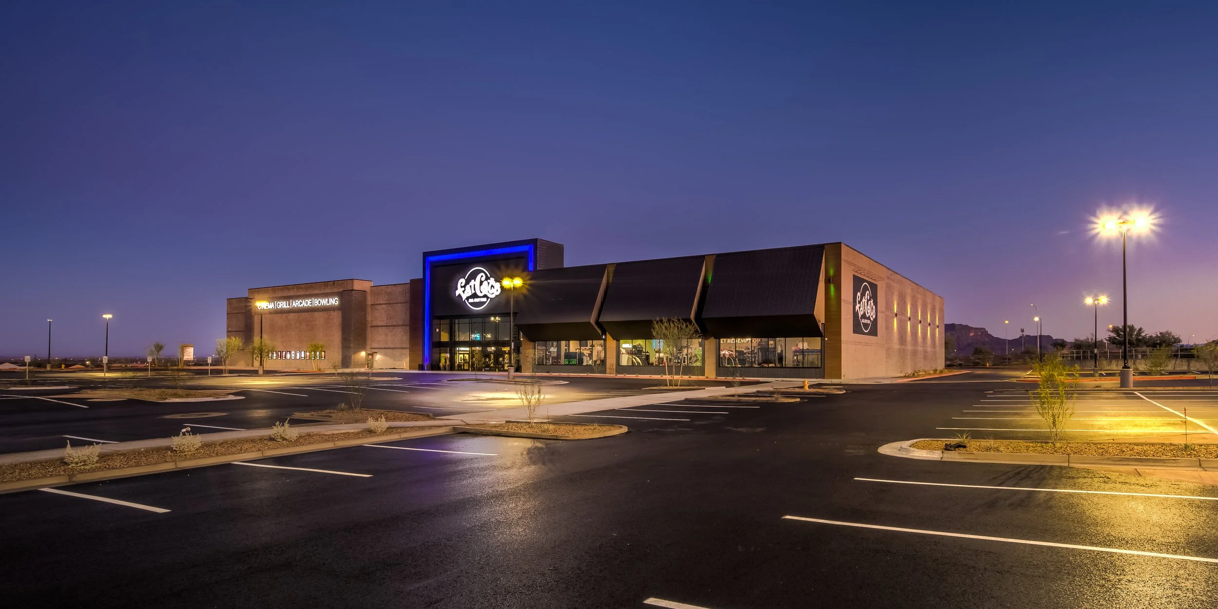 Large, modern building with signage for FatCats, a restaurant or entertainment venue, in a mostly empty parking lot during twilight with streetlights on and distant mountains in the background.