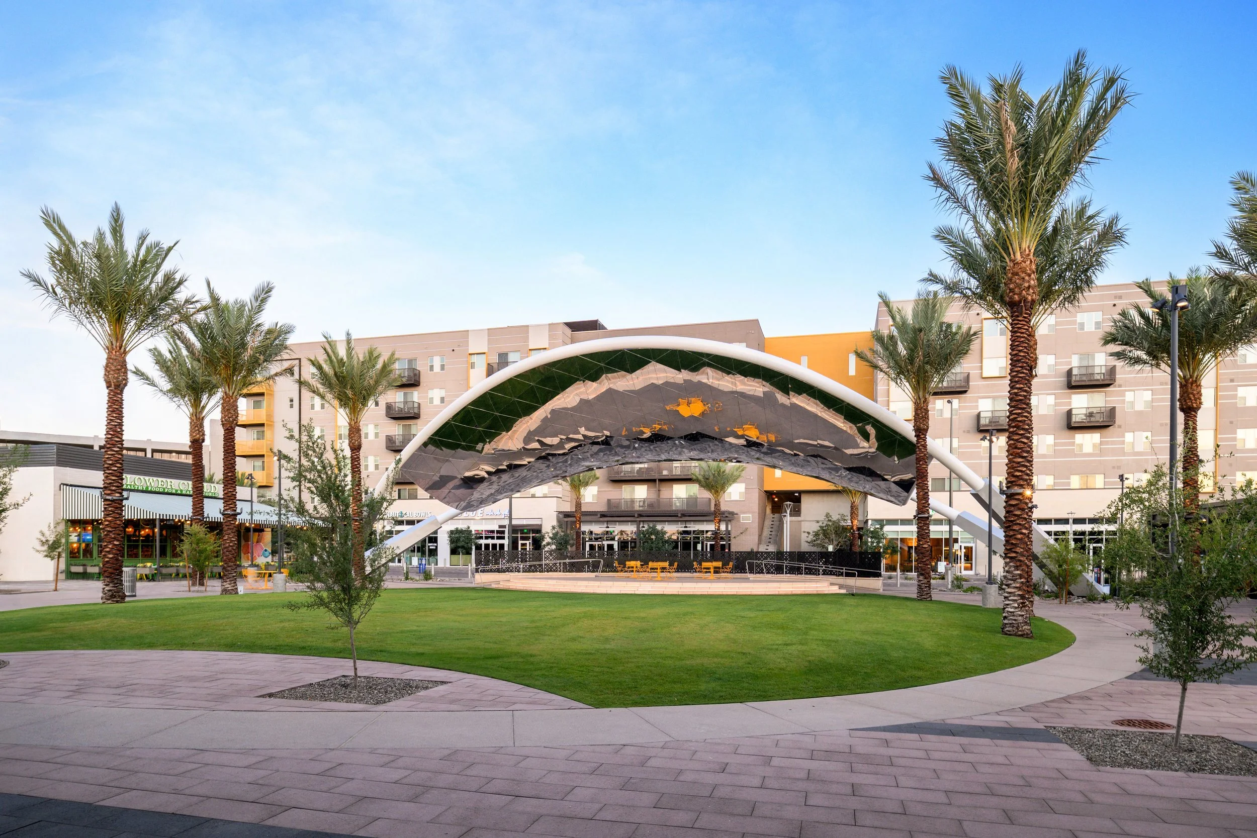 Public park with a stage under a large white arch, surrounded by palm trees and a grassy area, with multi-story apartment buildings in the background.