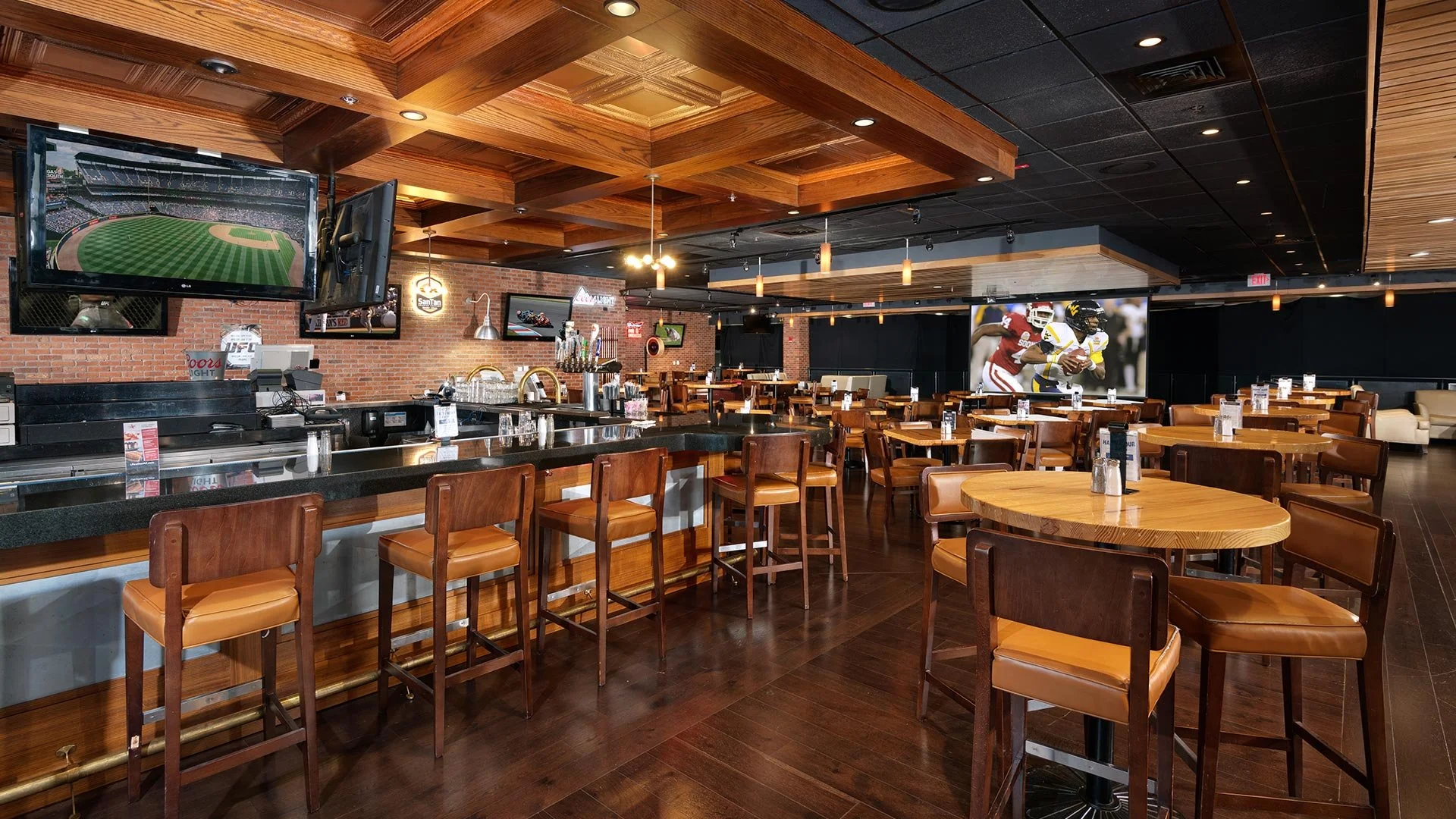 Interior of a sports bar with wooden ceiling, brick walls, multiple TVs displaying sports, a bar counter with seating, and tables and chairs for patrons.