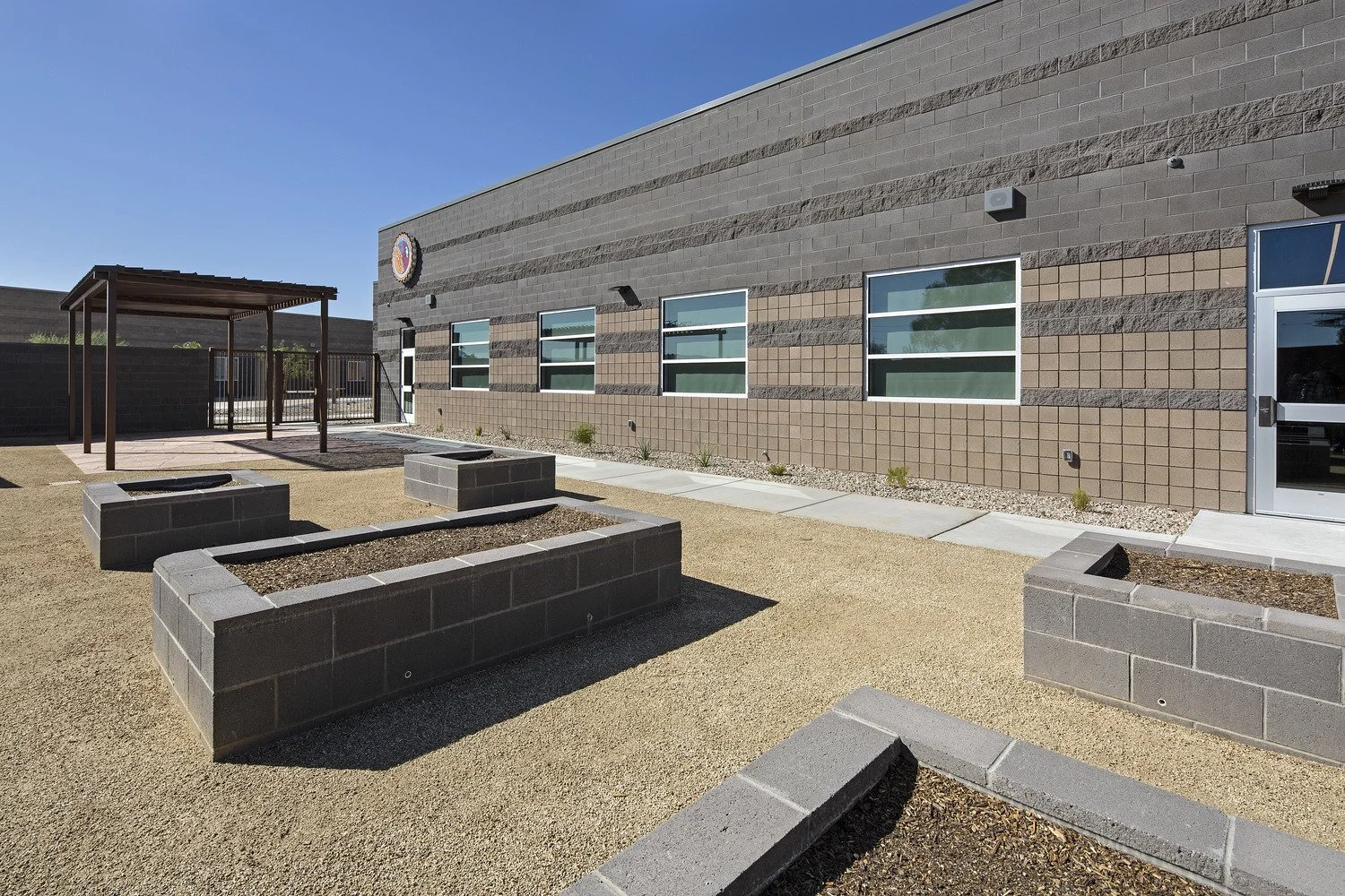 Exterior view of a modern school building with large windows, a small outside covered area, and empty brick planters in the courtyard under a clear blue sky.