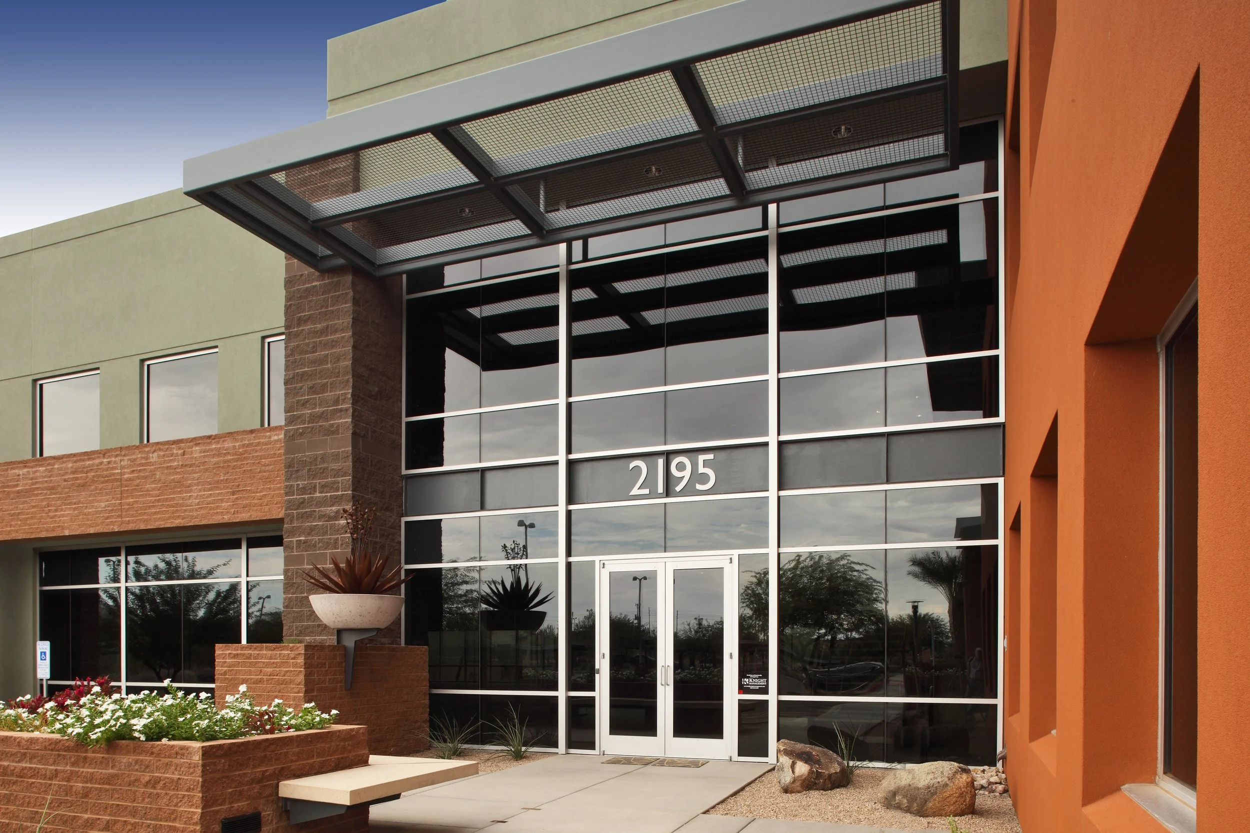 Modern commercial building with glass facade, the number 2195 displayed above the entrance, and a landscaped area with plants and rocks in front.