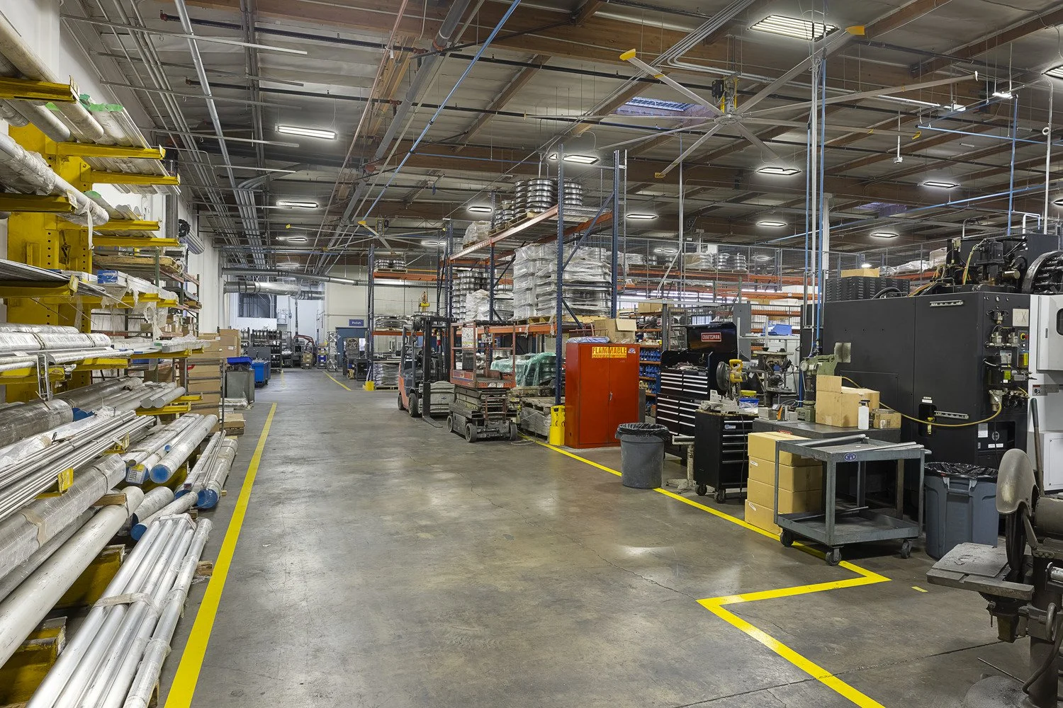 Inside an industrial warehouse or manufacturing facility with shelves and equipment, including pipes on the left, forklifts in the center, and machinery on the right, with yellow safety lines on the floor.