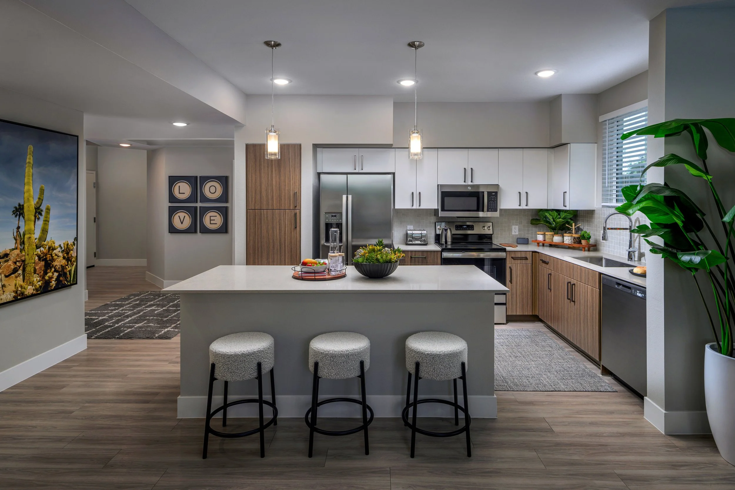 Modern kitchen with white and wood cabinets, stainless steel appliances, a white island with three stools, and decorative plants.