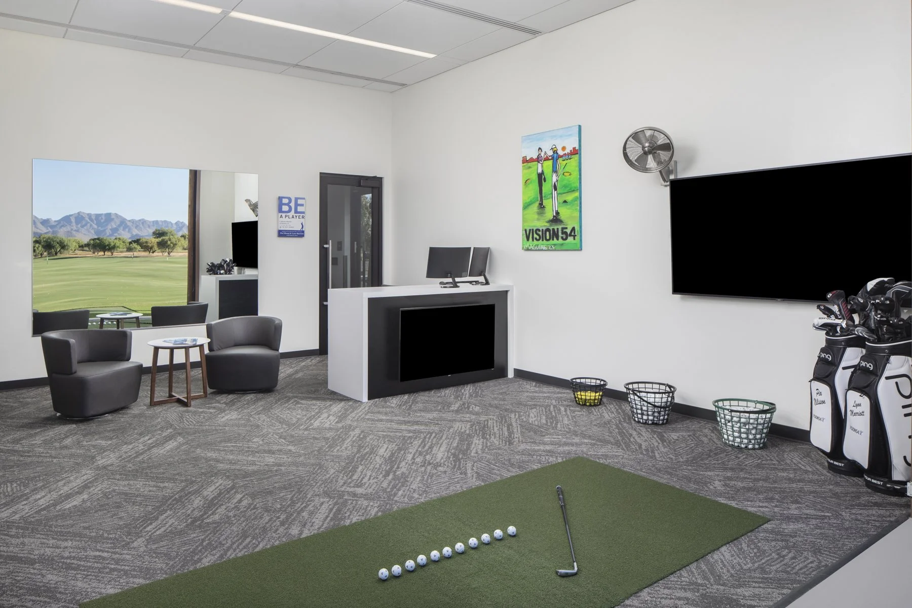 Indoor golf simulator room with a green turf, golf balls lined up, and a golf club. Modern furniture, a large flat-screen TV, and golf bags are in the room. A window shows an outdoor golf course and mountains.