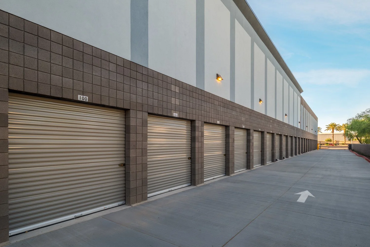 Exterior view of a storage facility with multiple closed beige roll-up doors, each numbered, situated along a clean concrete driveway with an arrow pointing forward, under a clear blue sky with some trees in the background.