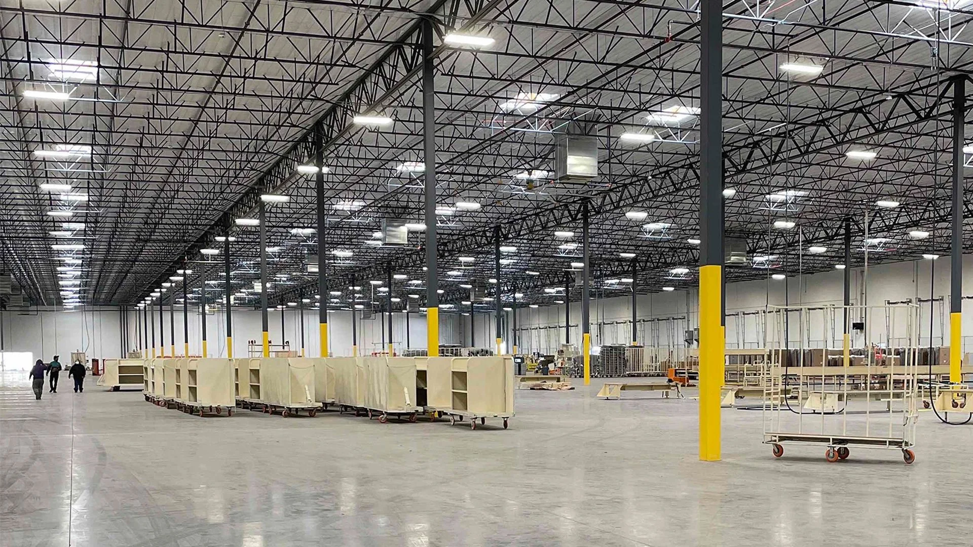 Inside a large, mostly empty warehouse or distribution center with a high ceiling, metal framework, and bright overhead lighting. Several empty carts and shelving units are scattered around the space, with three people walking in the distance.