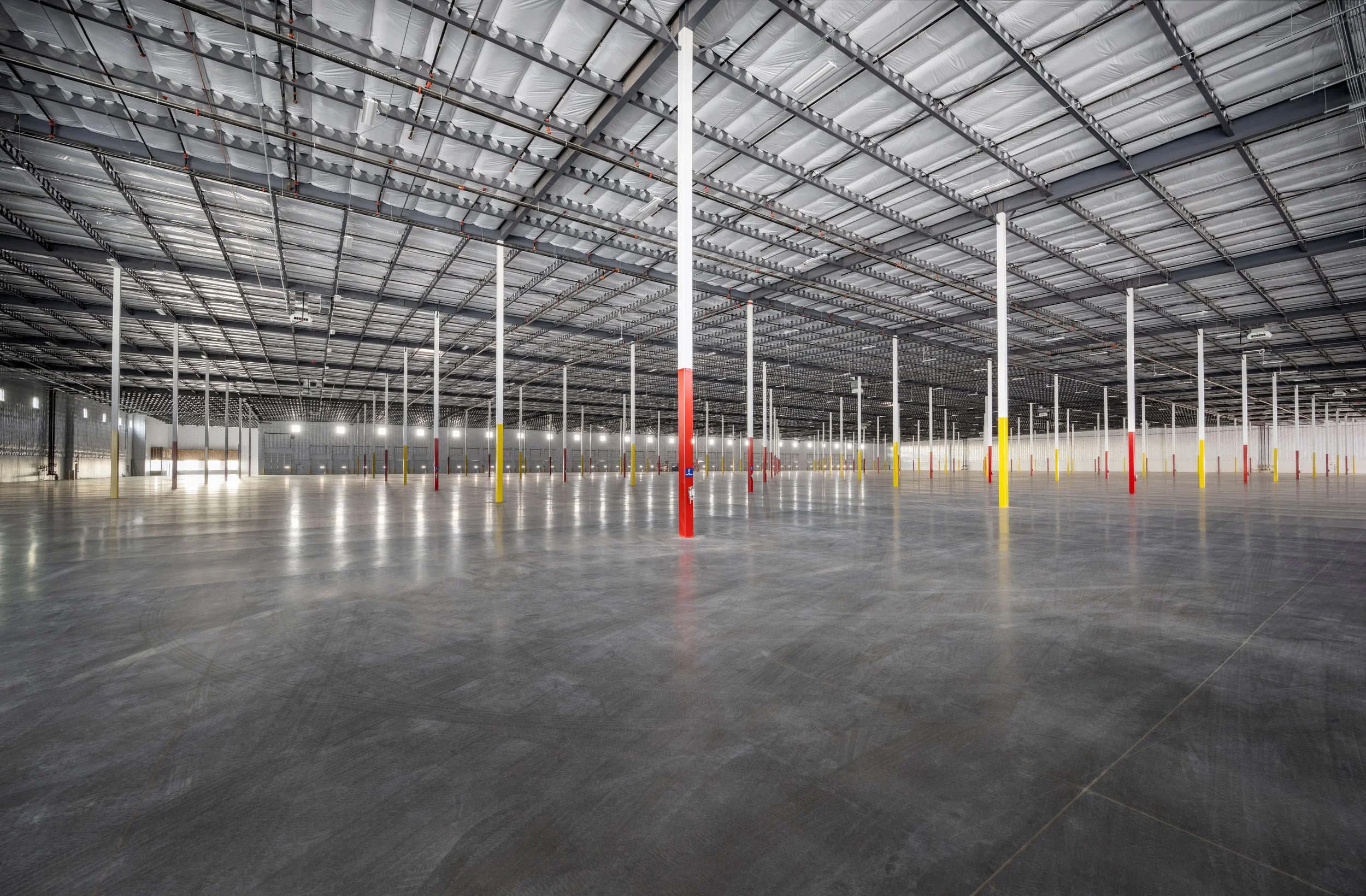 Empty warehouse with polished concrete floor, tall white support columns painted red and yellow, and a high metal ceiling with insulation and lighting fixtures.