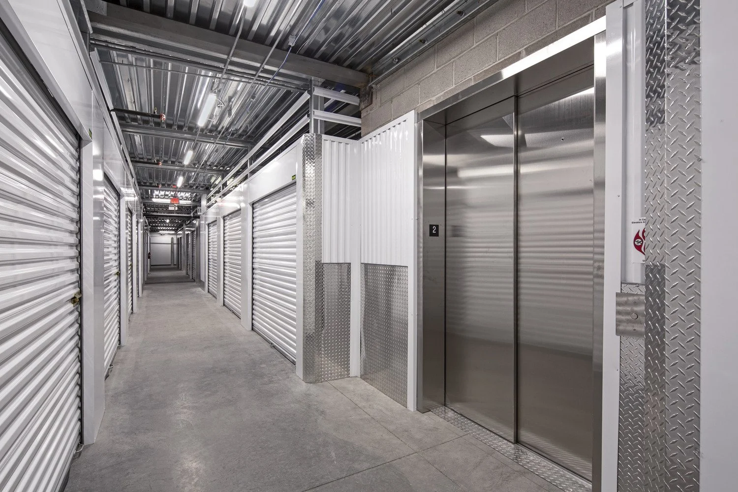 Industrial storage facility with metal roll-up doors, an elevator with stainless steel doors, concrete flooring, and exposed ceiling infrastructure.