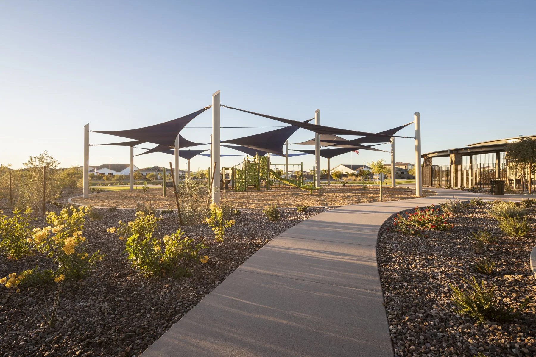 A playground with a shaded canopy, a paved walking path, and surrounding desert plants in a landscaped park area, with houses visible in the background during sunset.
