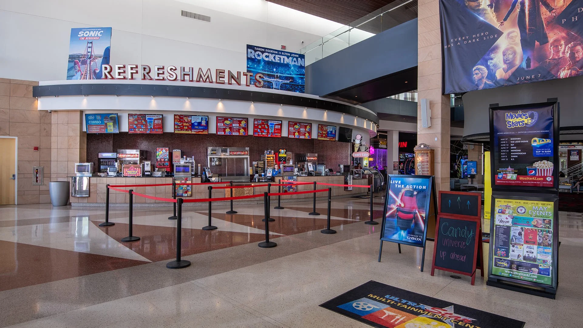 Movie theater concession stand with digital menu boards, surrounding posters, and signs advertising movies and snacks, with barriers guiding customers.