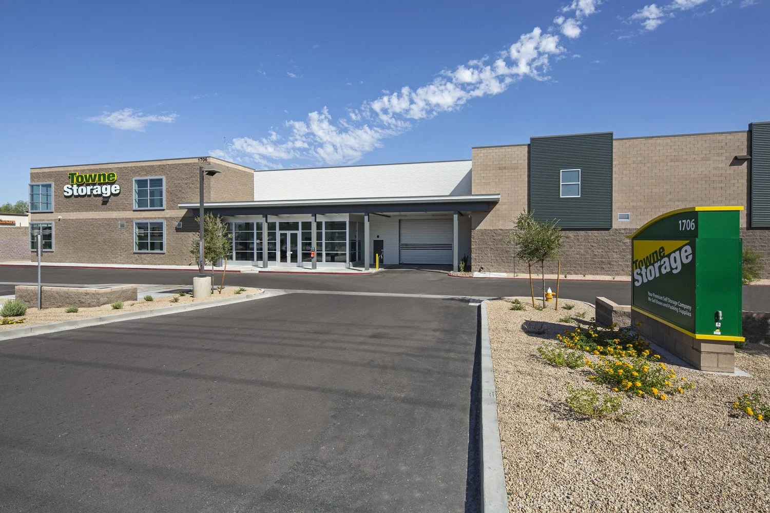 Exterior view of Towne Storage facility with a large parking area in front, small trees, and a landscaped area with flowers and gravel. The building has a modern design with brick and metal siding, and a prominent green and yellow storage sign with t
