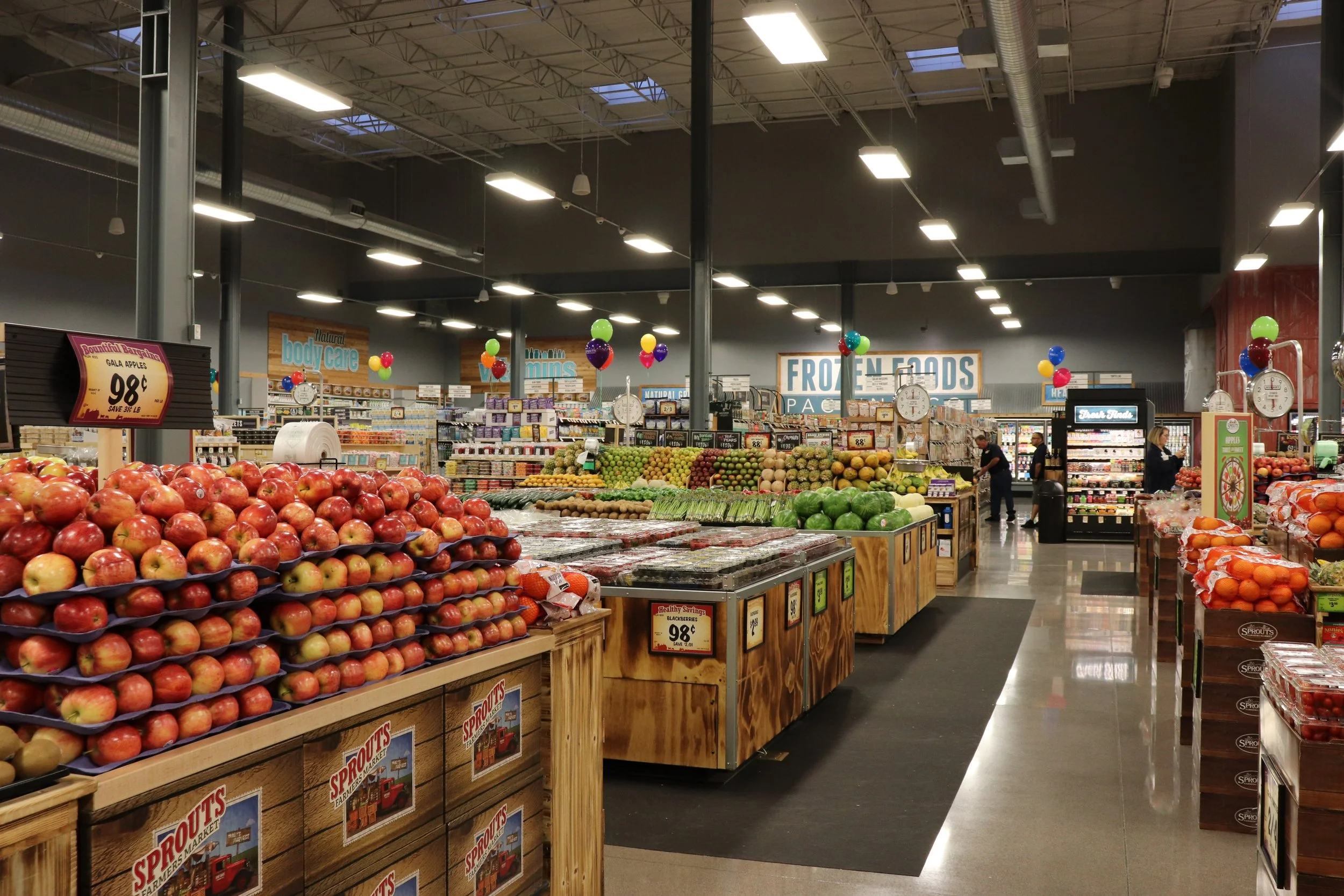 Interior of a grocery store displaying fresh apples, watermelons, oranges, and other produce, with signs for frozen foods and health care section, decorated with colorful balloons.