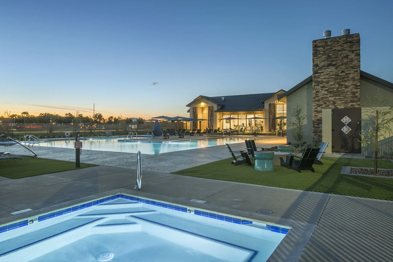 An outdoor swimming pool area at dusk, featuring a hot tub in the foreground and a larger pool with lounge chairs and umbrellas in the background, adjacent to a modern clubhouse with large windows and a brick chimney.