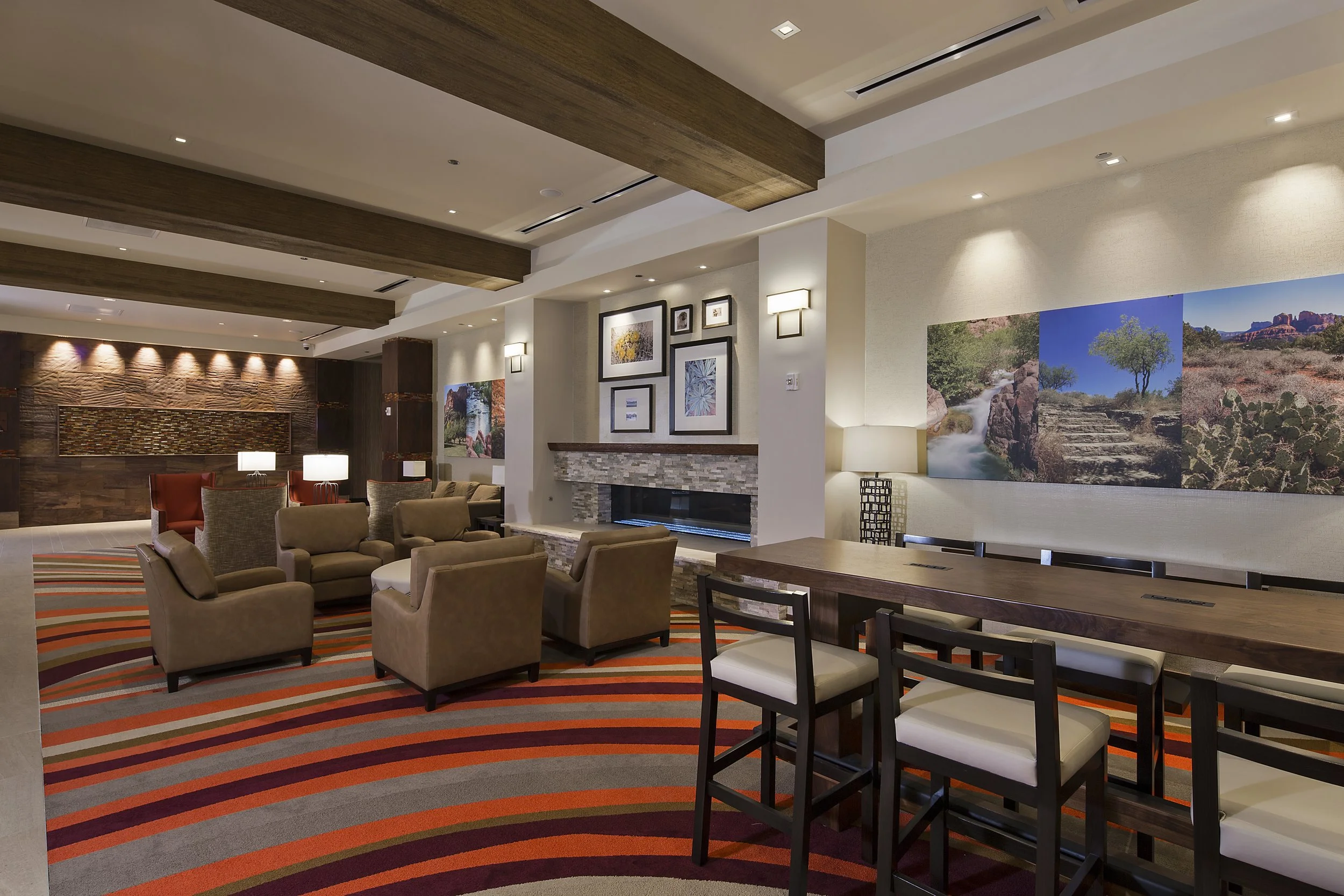 A cozy hotel lobby with beige chairs and red armchairs arranged for seating, a fireplace with framed art above it, colorful striped carpet, and large landscape photographs on the walls.