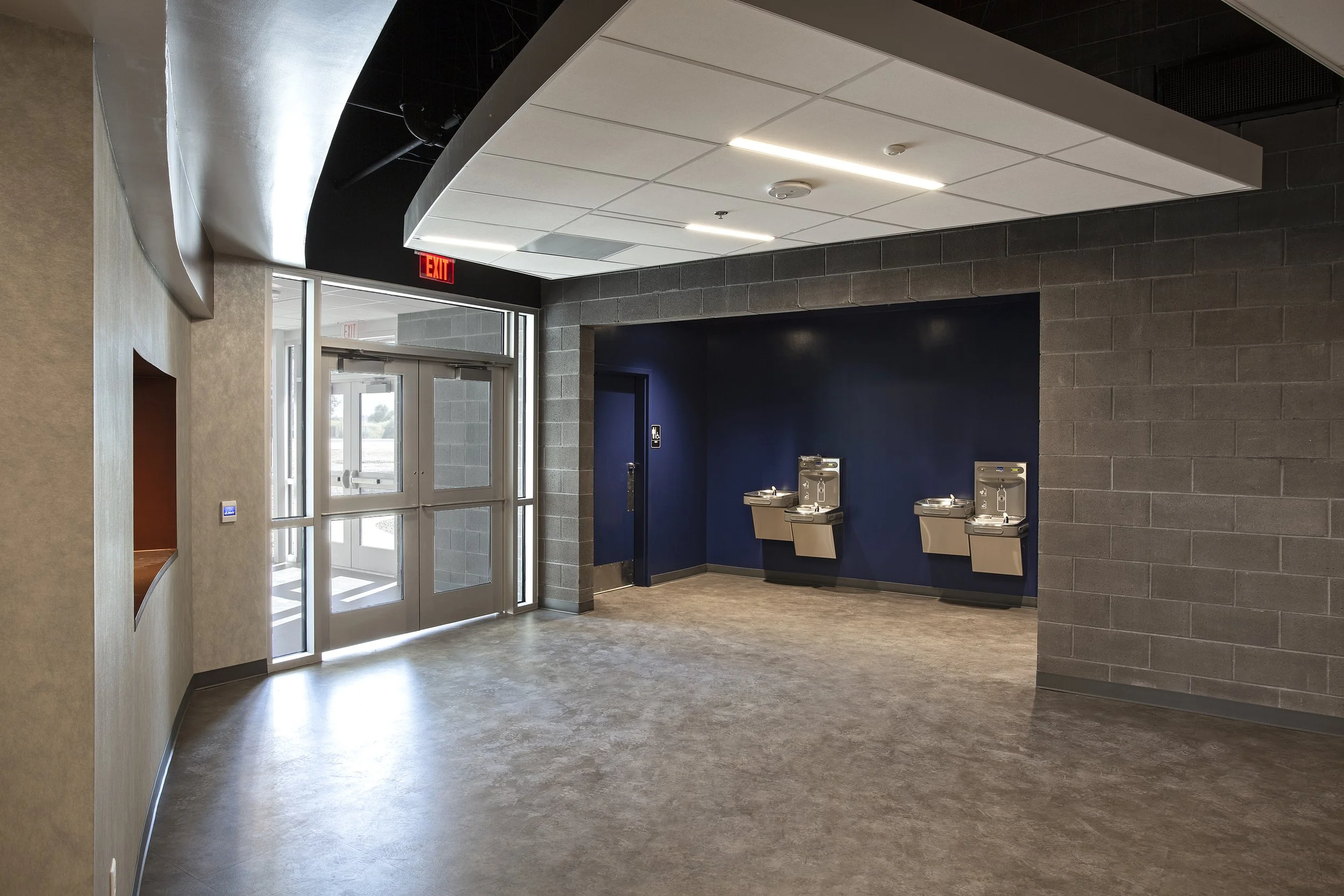 An indoor public space with a glass double door entrance, water fountains mounted on a dark blue wall, and an exit sign above the door.