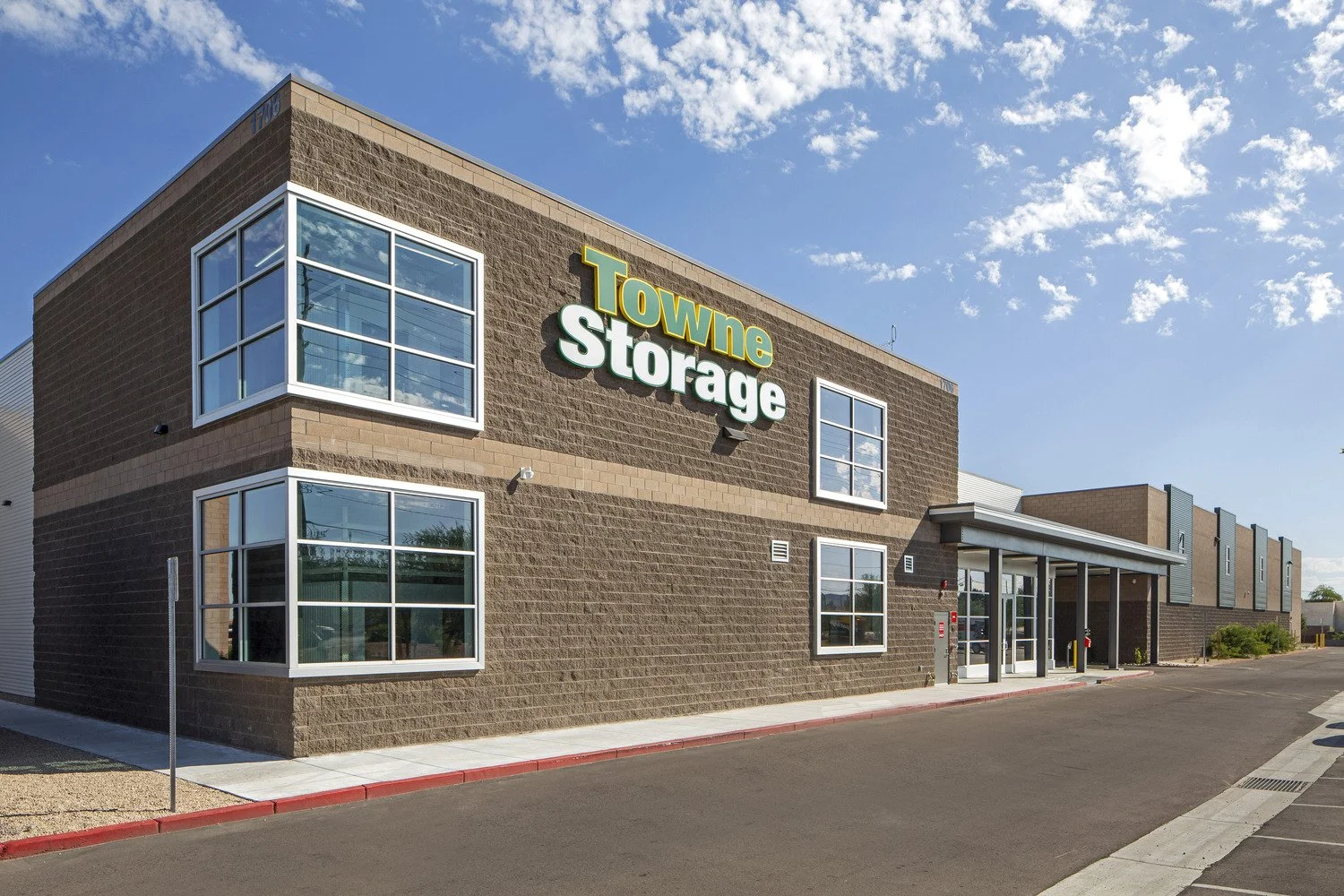 Exterior of a brick storage facility with large windows, a sign that reads 'Towne Storage,' and a parking lot in front on a sunny day.