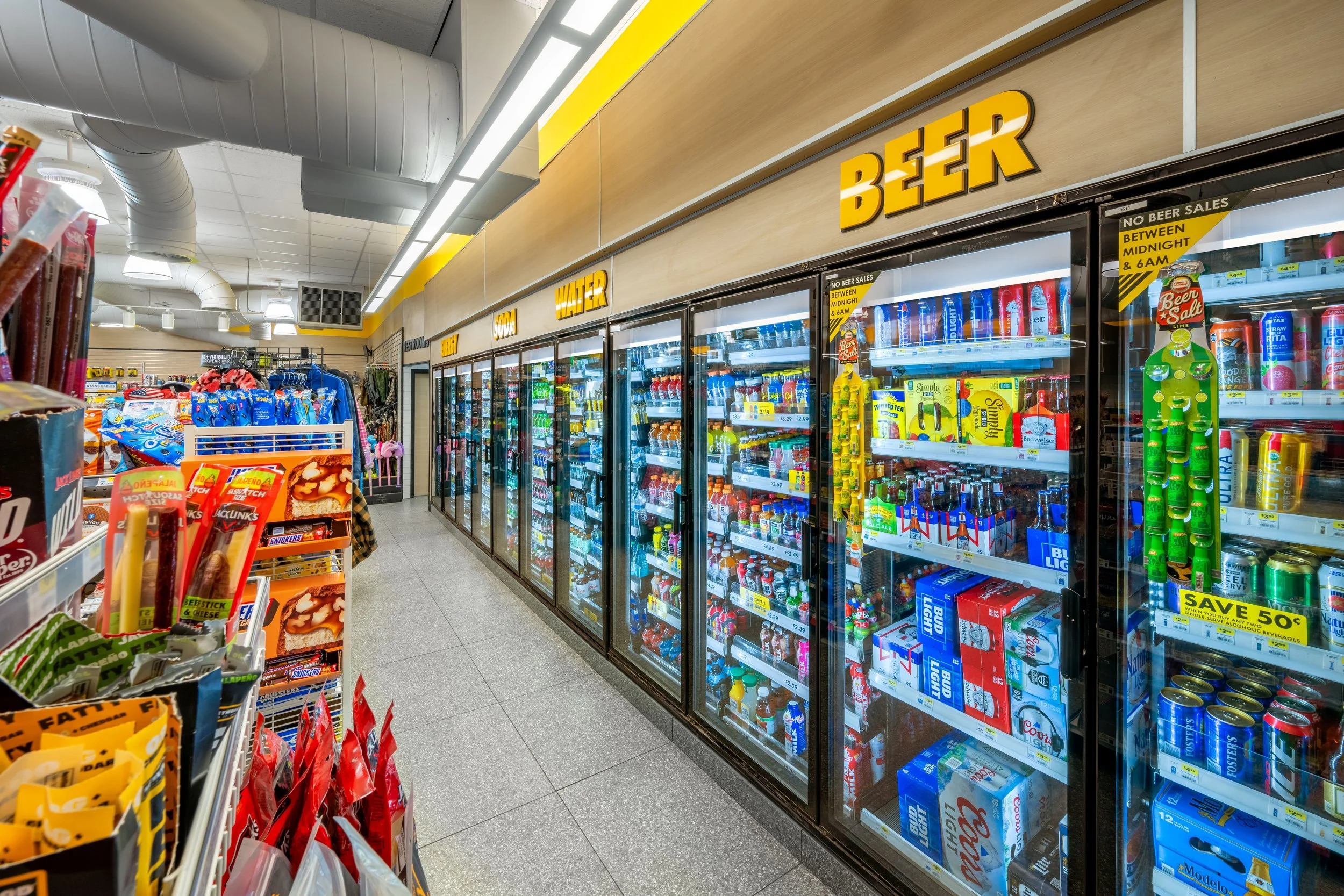 A convenience store aisle with drinks in refrigerated coolers labeled 'BEER.' The aisle also has snack shelves and other products.